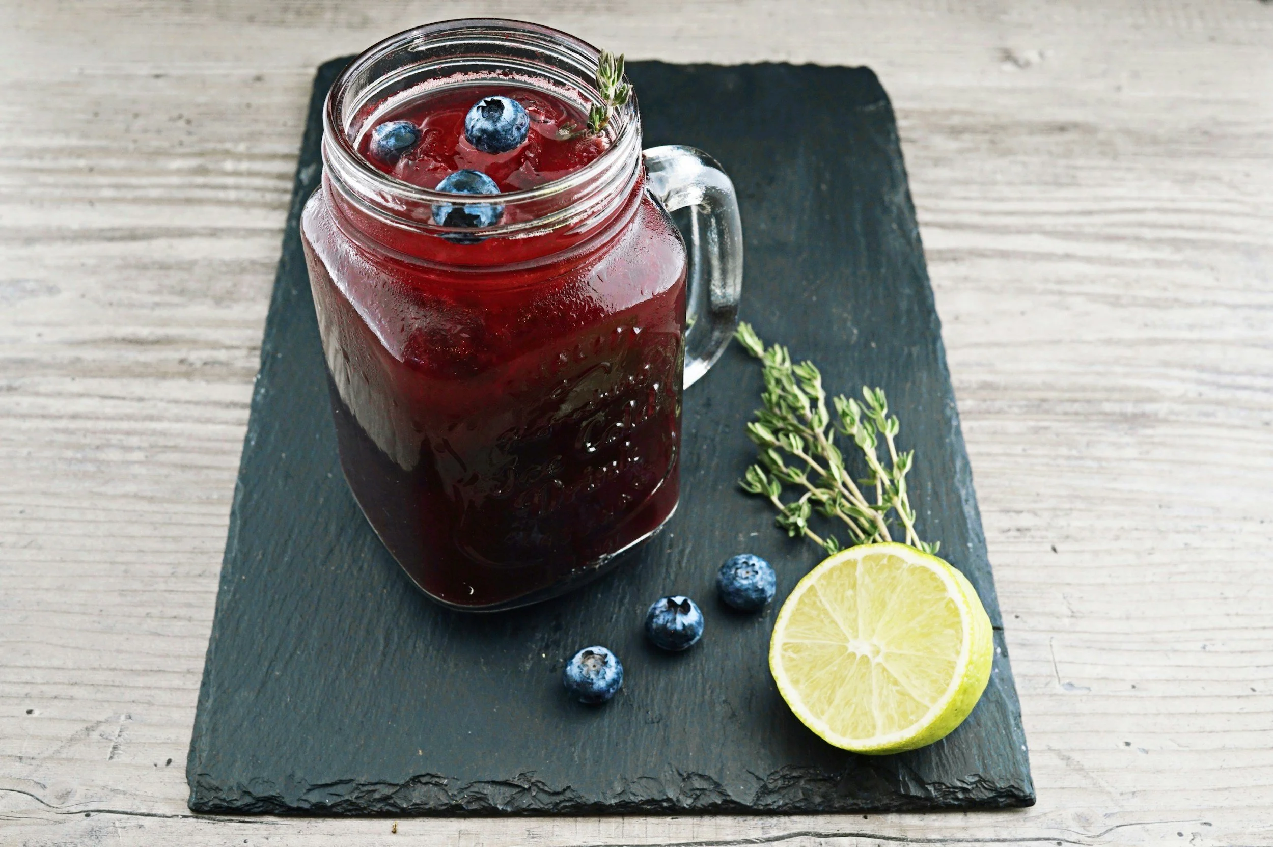 A glass mason jar with a handle filled with a dark red berry drink, garnished with blueberries and a sprig of thyme. The jar is placed on a black slate serving board alongside a halved lemon, a small bunch of thyme, and a few blueberries on a light wooden surface.