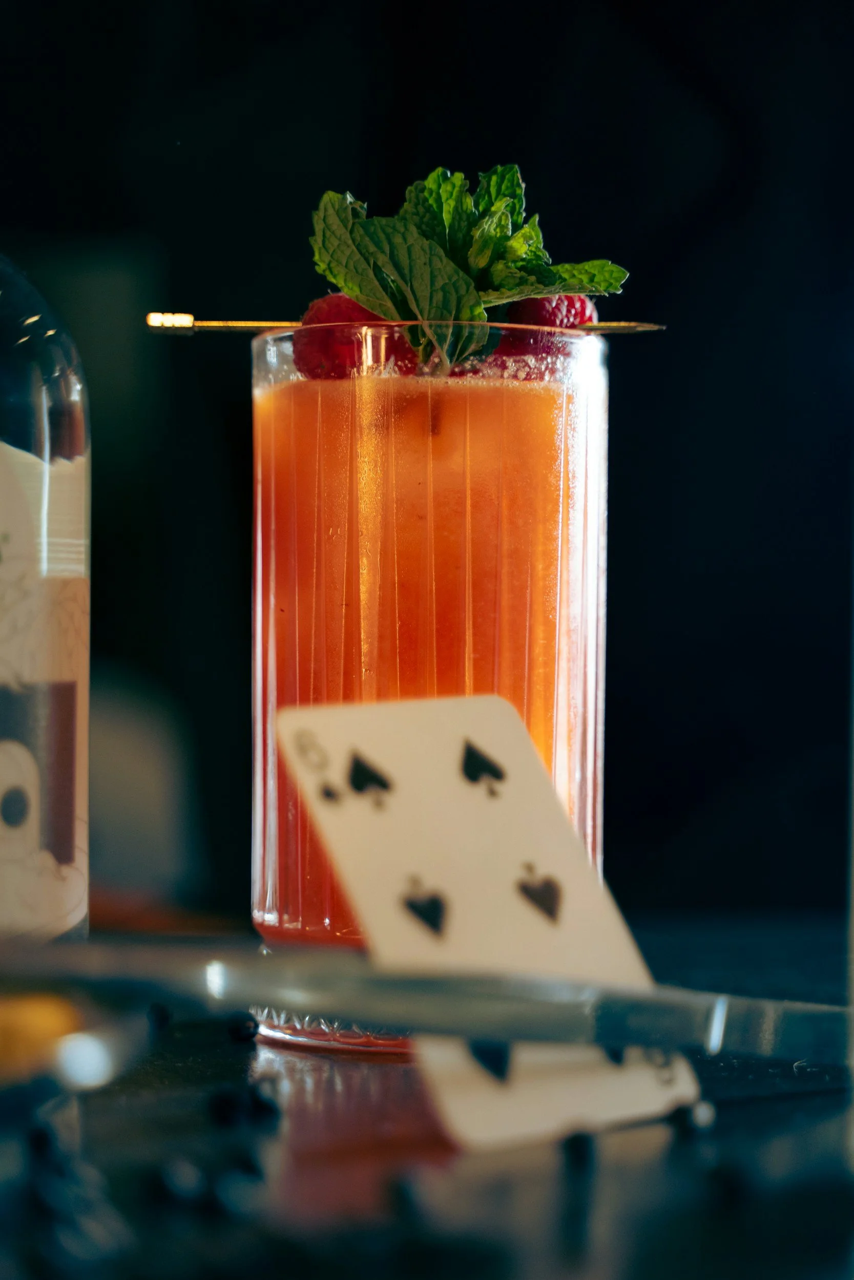 A tall glass of pink or orange cocktail garnished with fresh mint leaves and raspberries, with a playing card displaying four aces leaning against the glass.