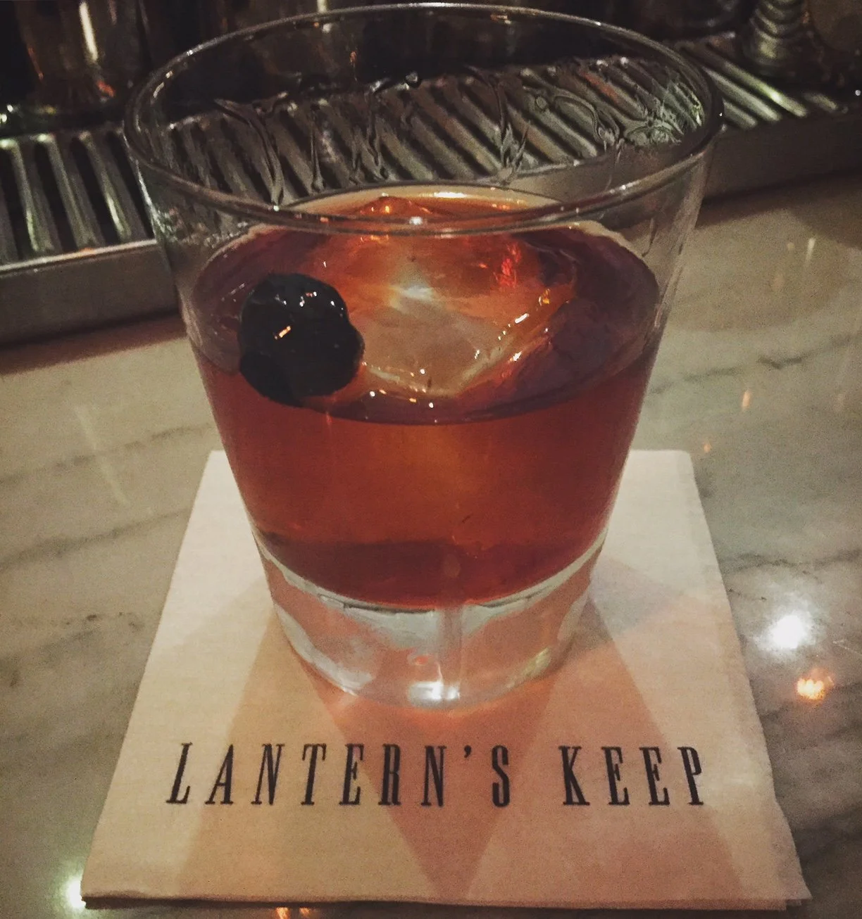 A cocktail with a cherry and an ice cube in a glass, placed on a napkin that reads 'LANTERN'S KEEP,' on a marble countertop.
