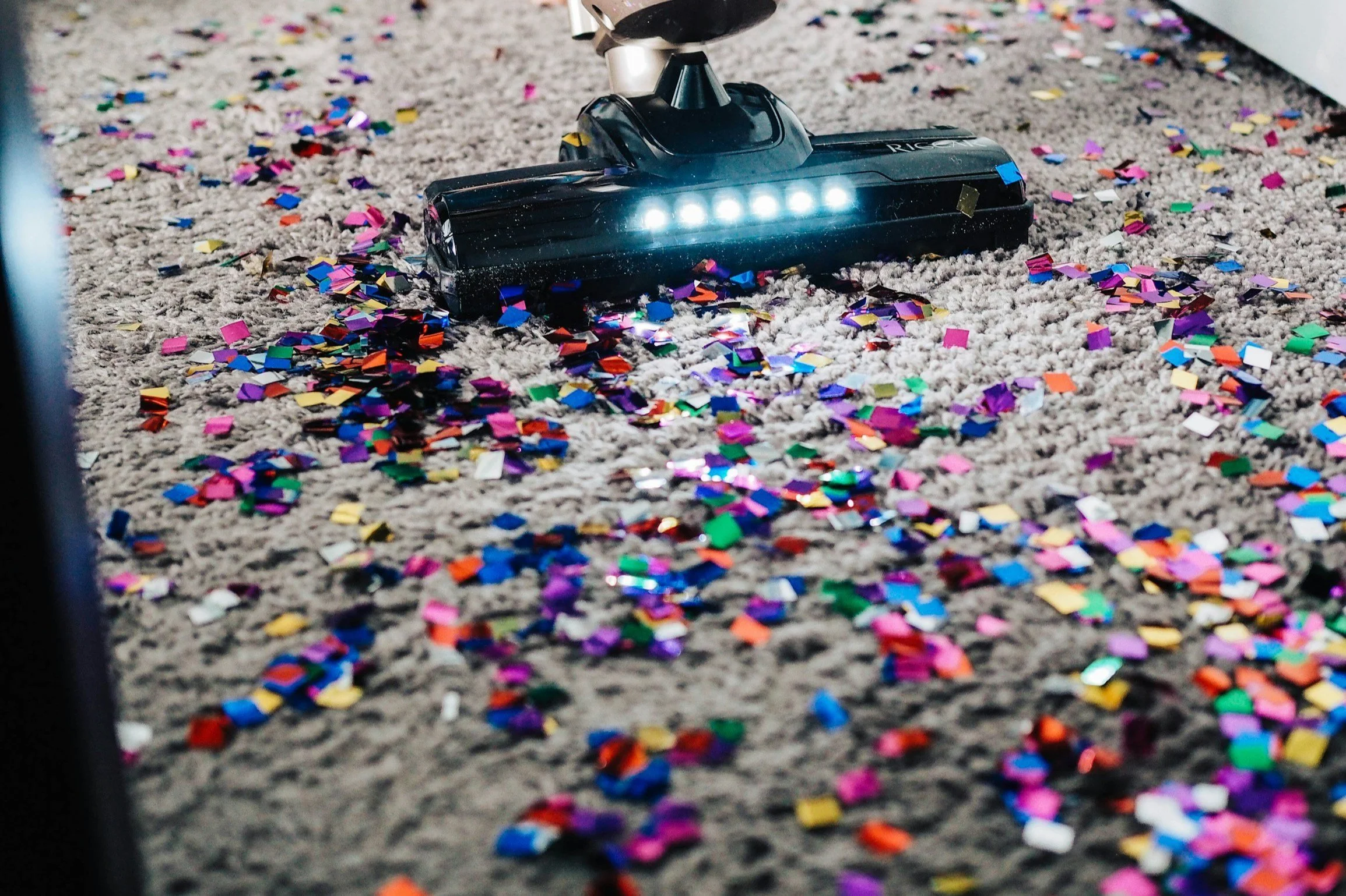 A vacuum cleaner with a light bar on it cleaning a beige carpet scattered with colorful confetti.