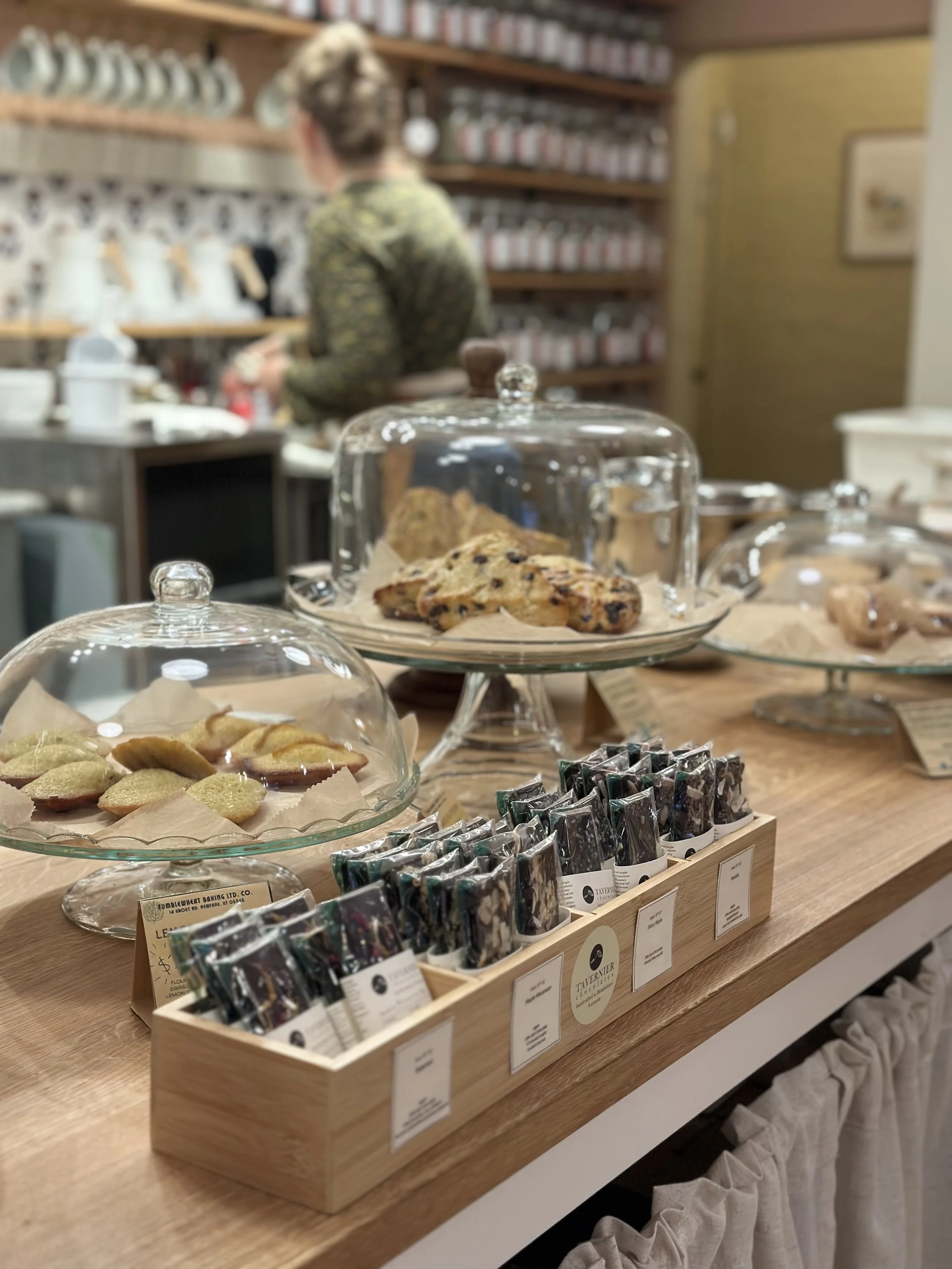 Assorted cookies and baked goods displayed under glass domes on a wooden counter in The Humble Mug, with a blurred background of a person browsing shelves.