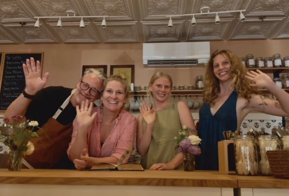 Four people of different ages smiling and waving behind a wooden counter in a cozy shop or cafe, with jars and flowers on the counter and a blackboard menu in the background.