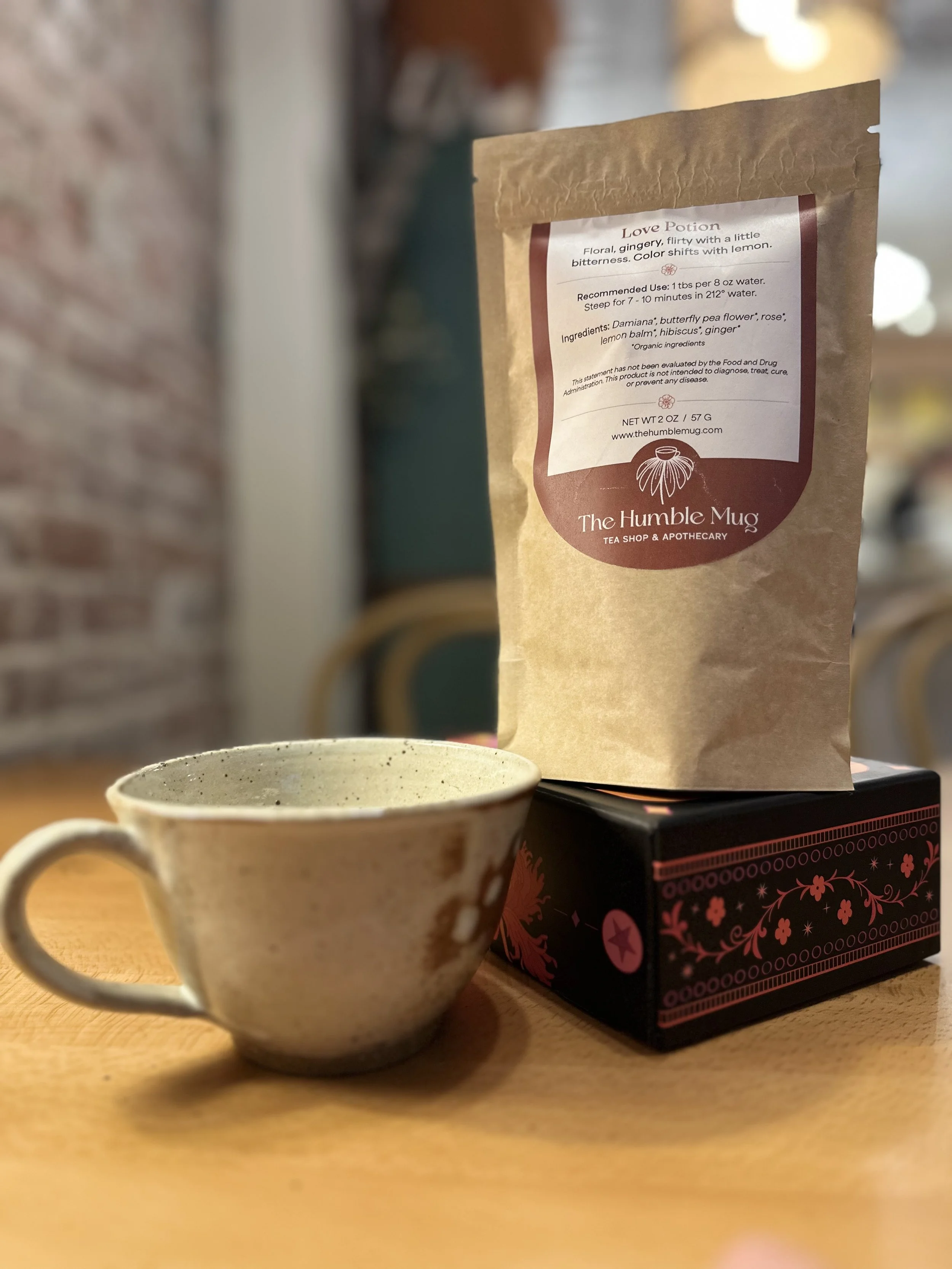 A ceramic mug filled with tea, a brown paper bag labeled 'Love Potion' from The Humble Mug, and a decorative black box with pink floral and star patterns on a wooden table.