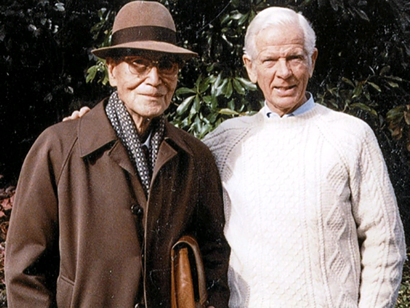Two elderly men standing outdoors in front of green foliage, one wearing a brown coat, hat, and scarf, the other in a white cable-knit sweater.