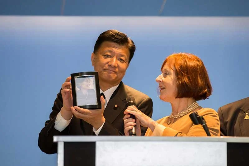 A man in a suit holding a tablet displaying a photo of a woman, standing next to a woman with red hair speaking into a microphone at a formal event.