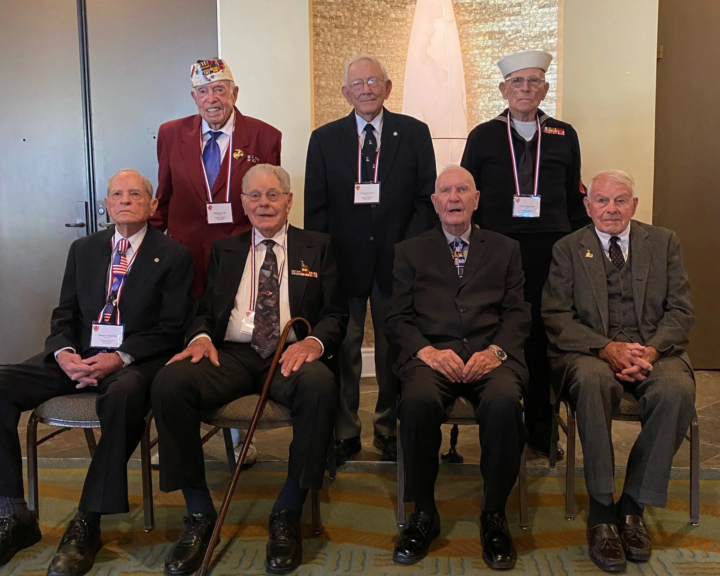 Group of seven elderly men, most wearing suits, some with hats, seated and standing in a professional setting, with one man on the right wearing a cap and a light blue shirt with a red lanyard.