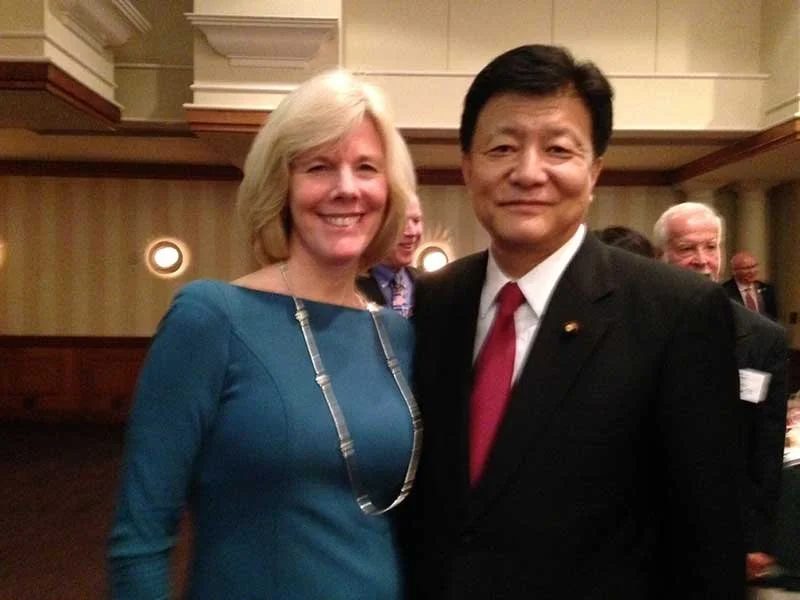 A woman with blonde hair wearing a blue dress and necklace, standing next to a man with black hair in a black suit, white shirt, and red tie at an indoor event.