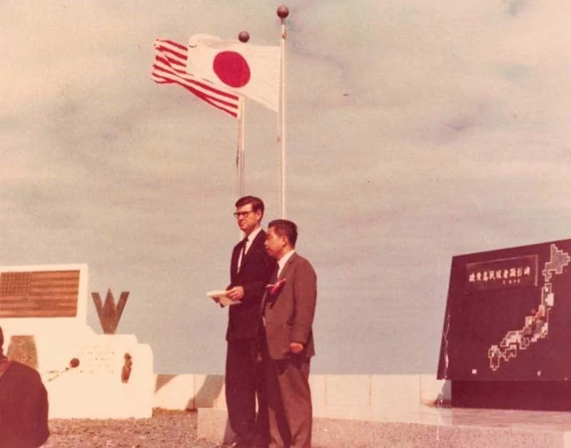Two men in suits standing outdoors, near Japanese and American flags. An informational board and large letter 'W' are visible in the background.