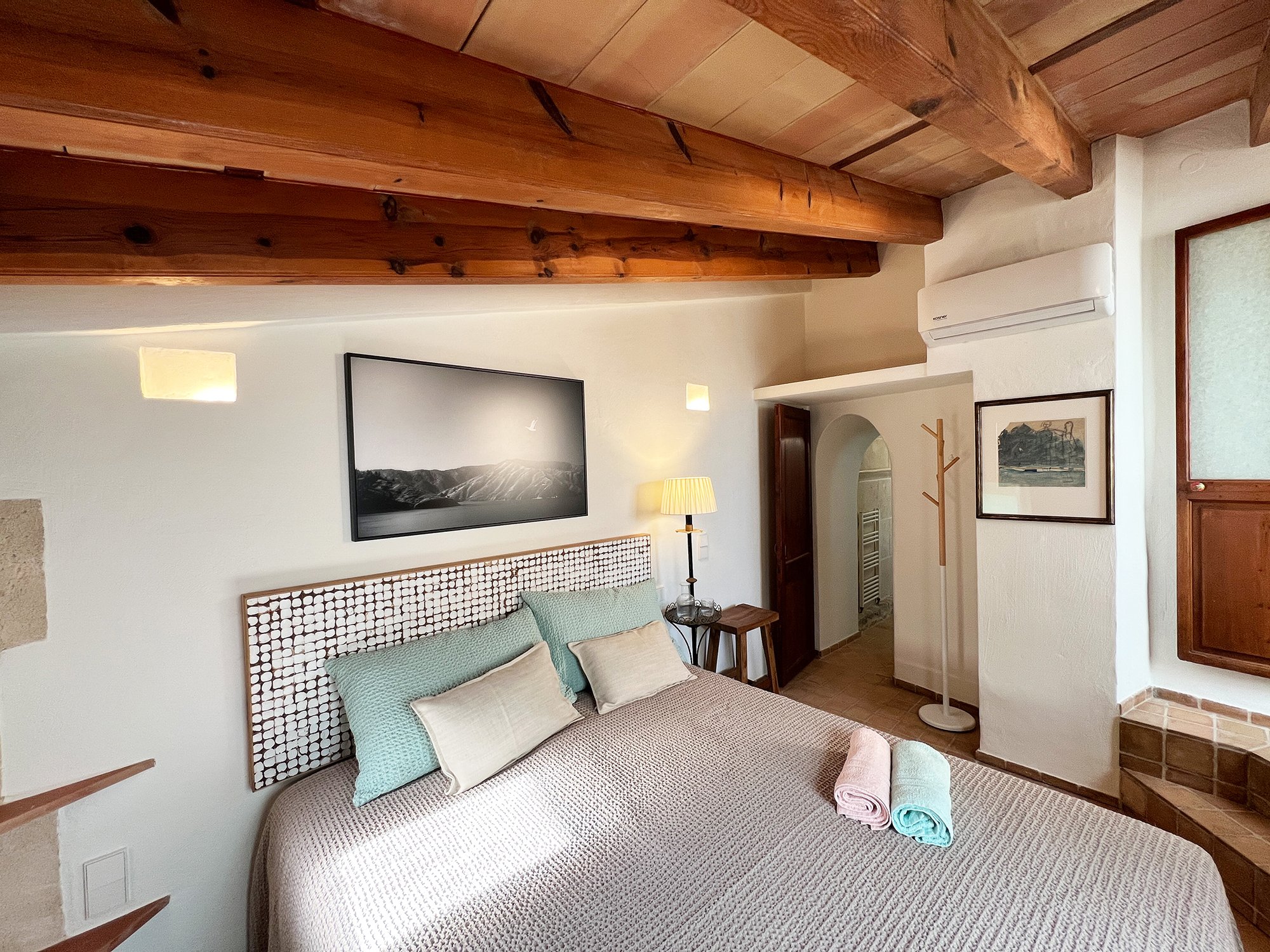 Gemütliches Schlafzimmer mit Holzbalken an der Decke, einem großen Bett mit dekorativen Kissen, einem Wandbild, einer Stehlampe und einem handtuchgekrönten Tisch.