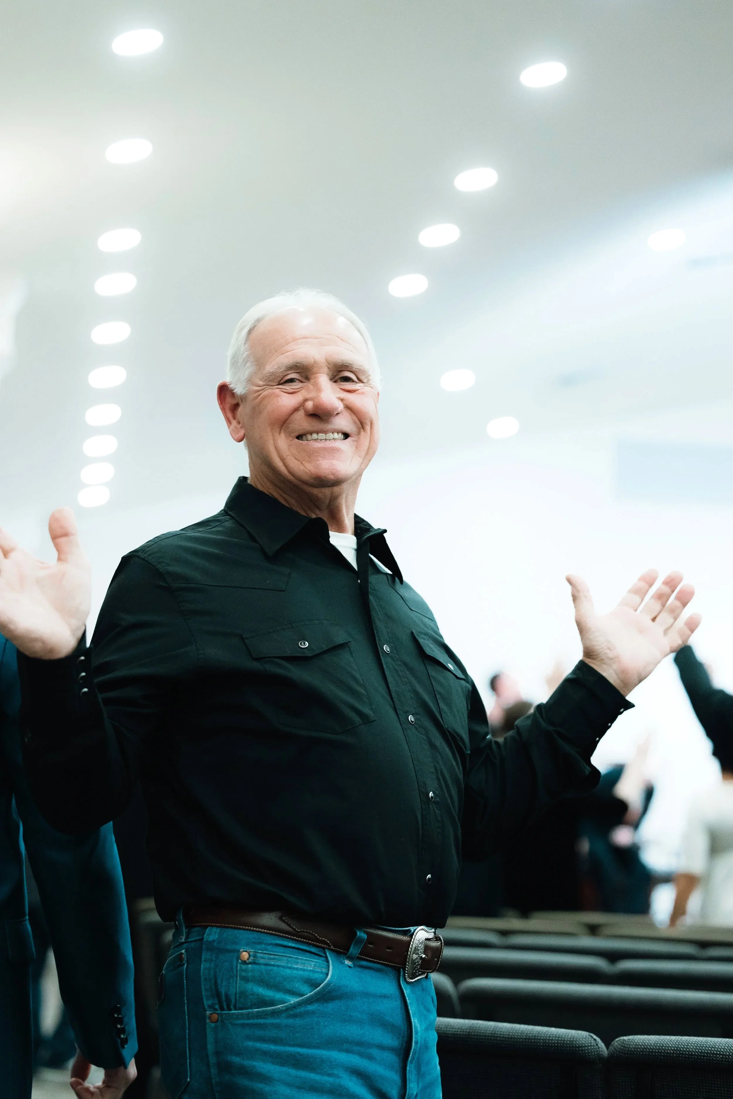 An elderly man with gray hair smiling and raising his hands in a welcoming gesture at an indoor event or gathering.