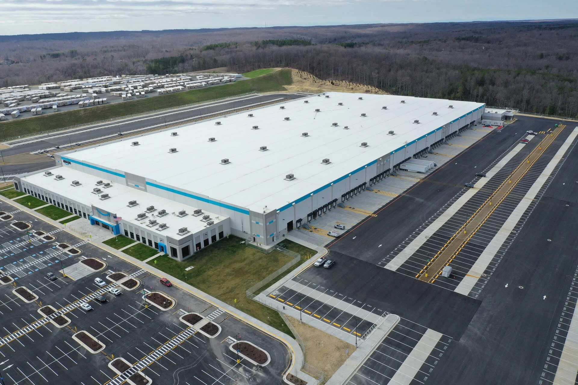 Aerial view of a large Amazon distribution warehouse with loading docks, parking lot, and surrounding forest.