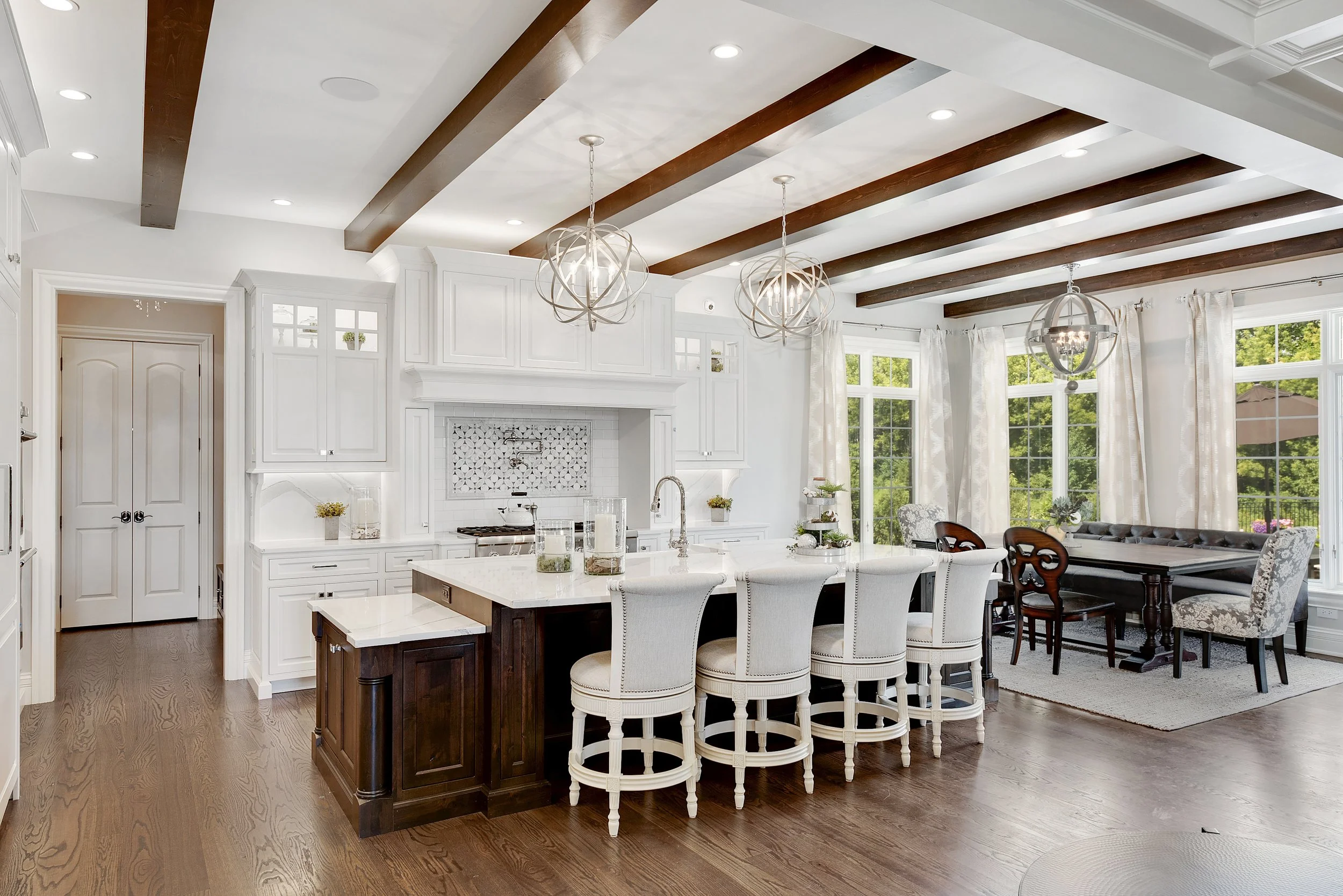 Bright kitchen and dining area with white cabinetry, a dark wood island, white bar stools, and a dining table with upholstered chairs. Large windows with white curtains let in natural light, and decorative light fixtures hang from a ceiling with dark wooden beams.