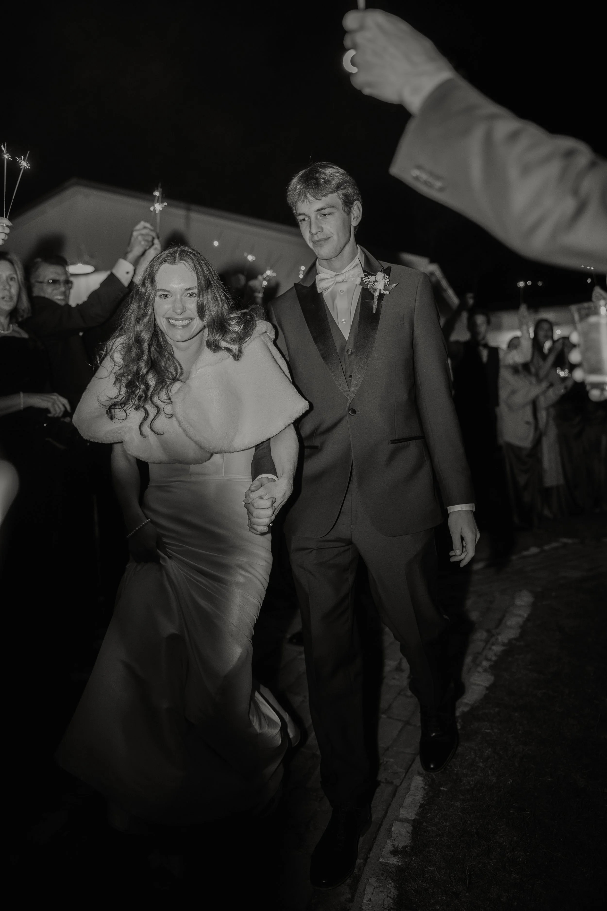 Black and white photo of a smiling woman and a man walking together at a nighttime celebration, surrounded by people holding sparklers.