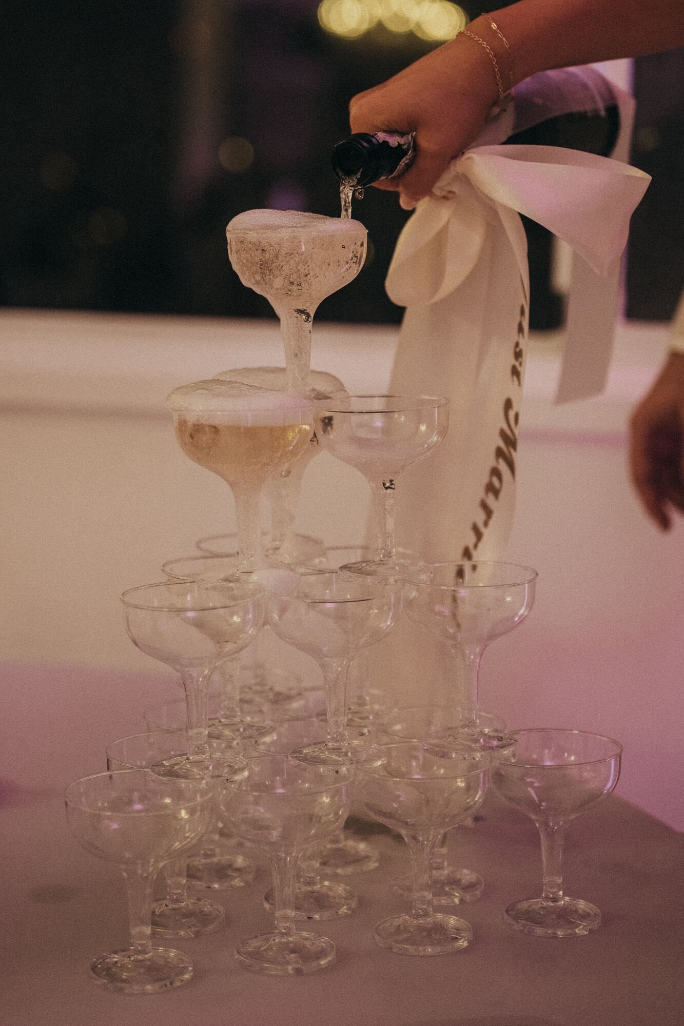 A person is pouring liquid from a bottle into a champagne glass on top of a pyramid of empty glasses.