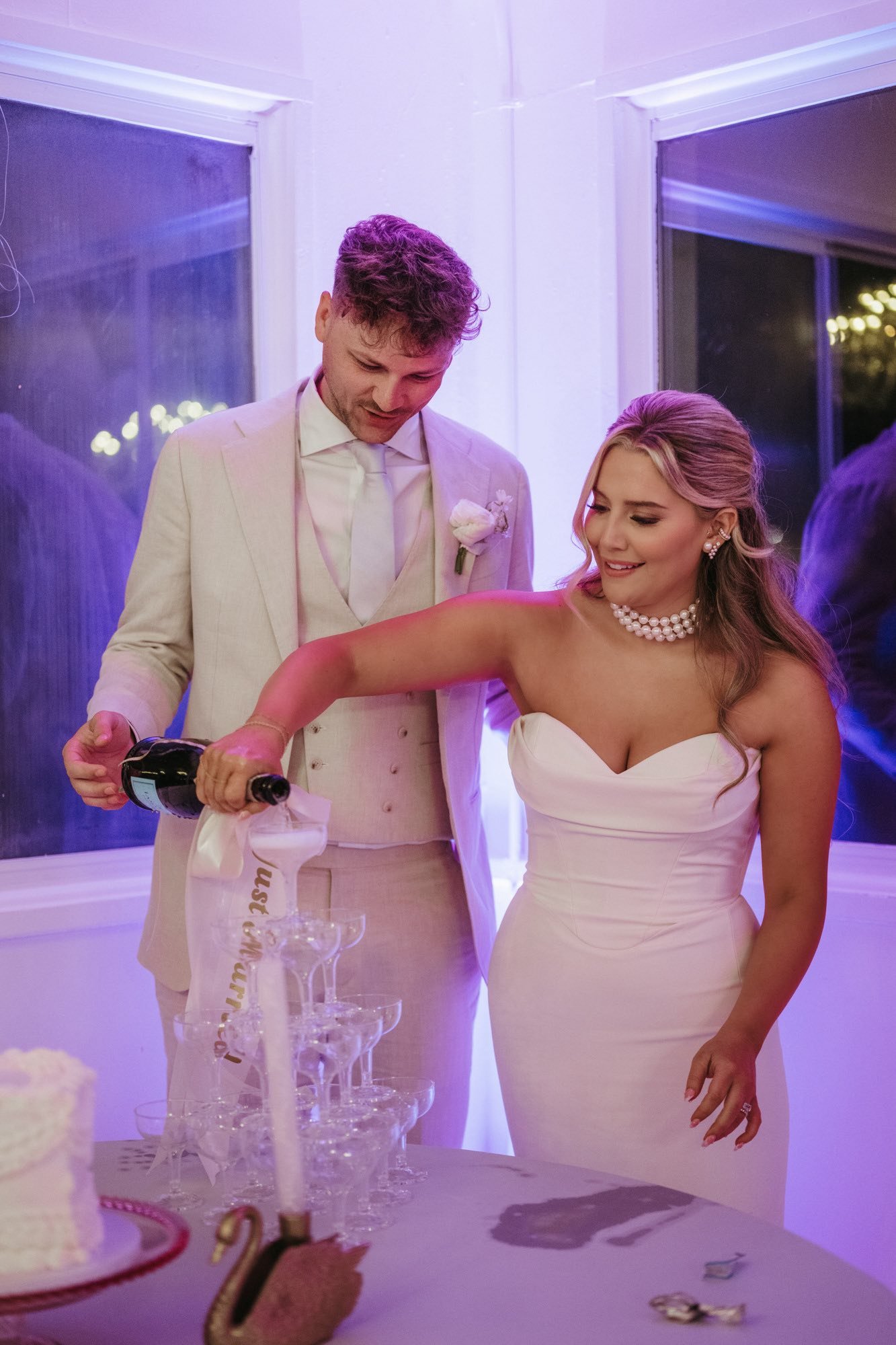 A newlywed couple pours champagne into glasses at their wedding reception, with the bride dressed in a strapless white wedding gown and the groom in a cream-colored suit.