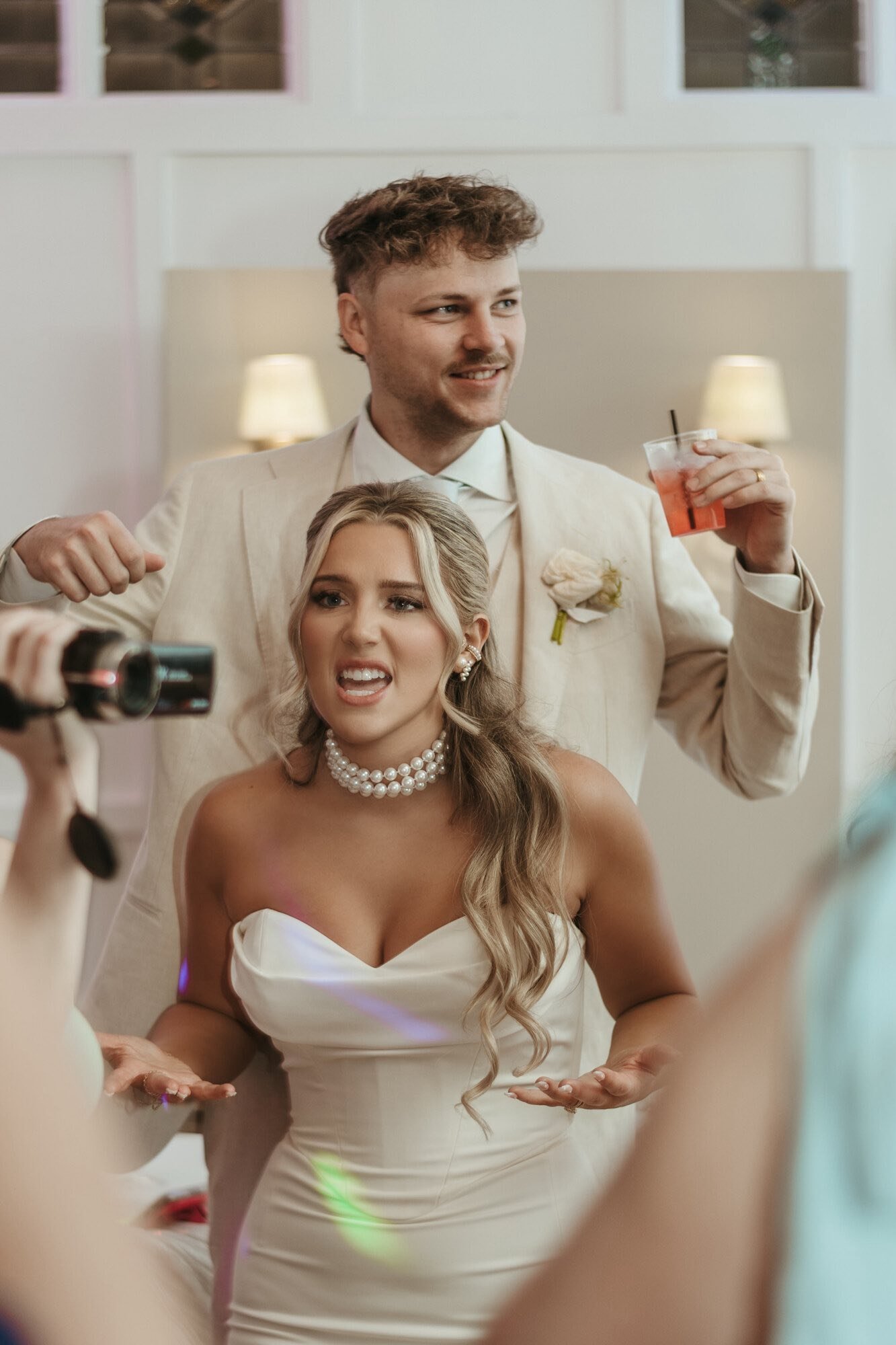 A bride in a white strapless dress with pearl jewelry and a man in a beige suit at a wedding. The man is holding a drink and smiling, while the bride appears surprised or confused as someone records them.