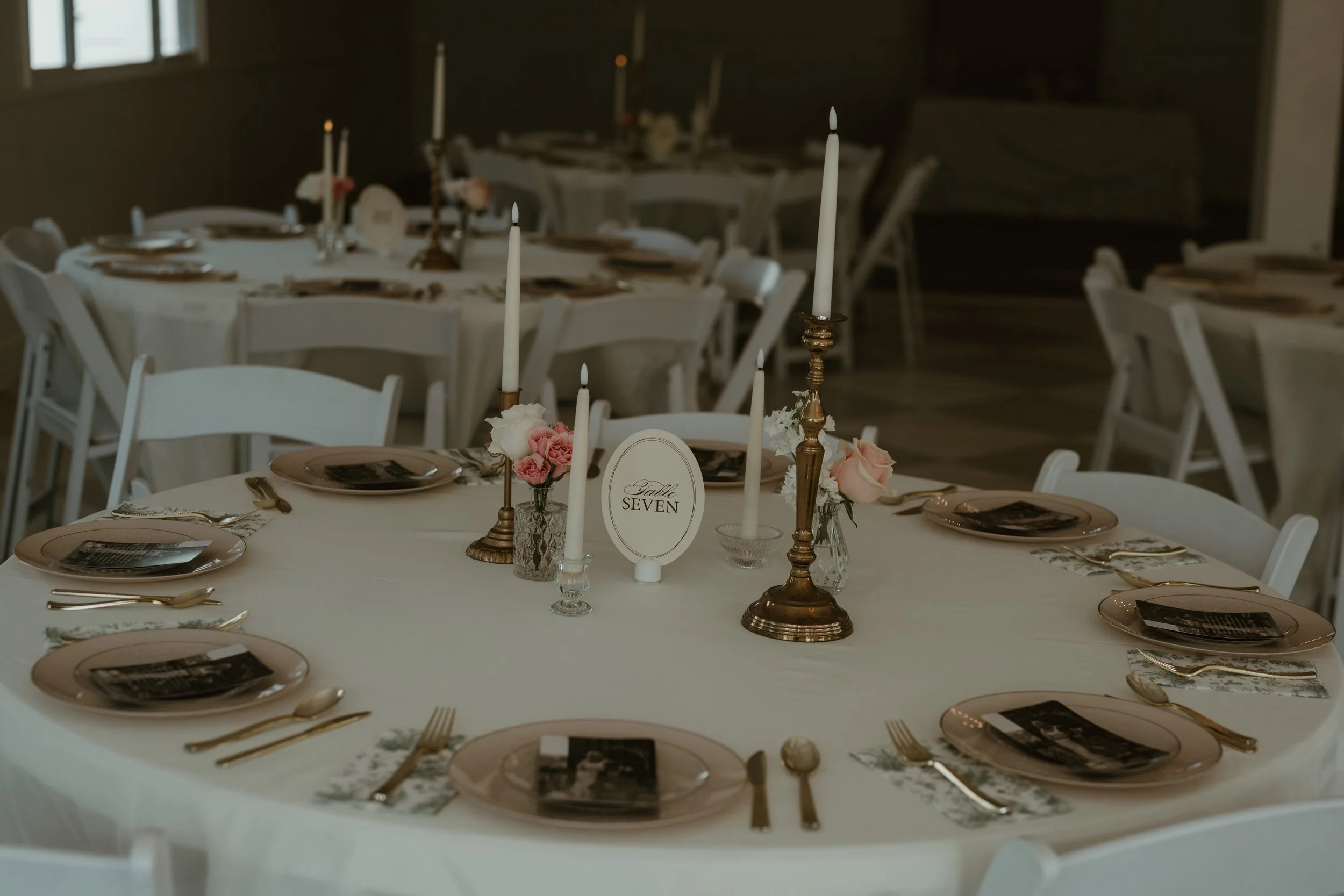 Round table set for a formal event with white tablecloth, plates, gold utensils, floral centerpieces, and tall candles, with chairs arranged around it.