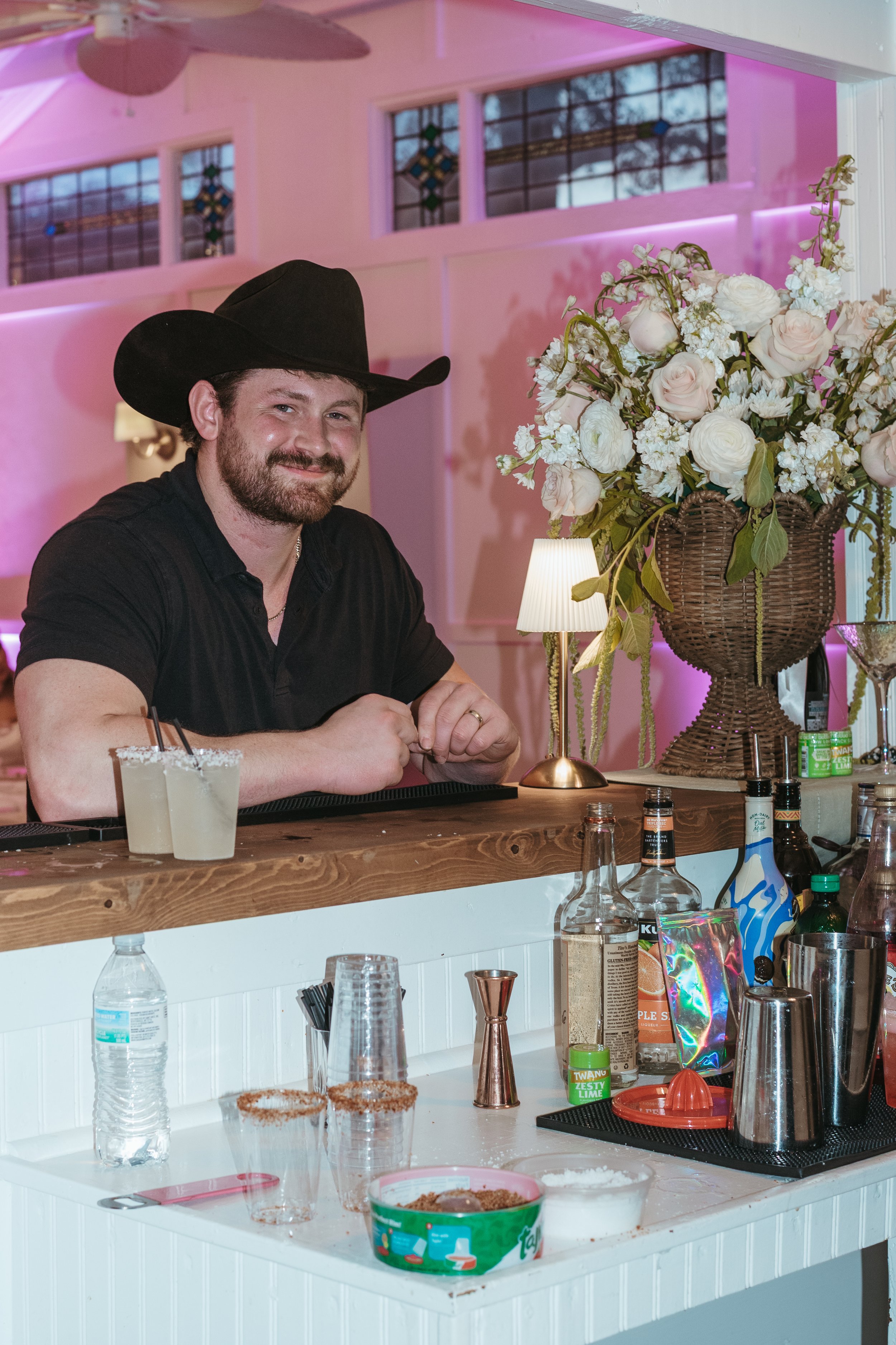 A man wearing a black cowboy hat and black shirt sitting at a bar counter decorated with a large floral arrangement and small table lamps. The bar has various bottles, glasses, and bartending tools, with a pink and purple lighting ambiance in the bac