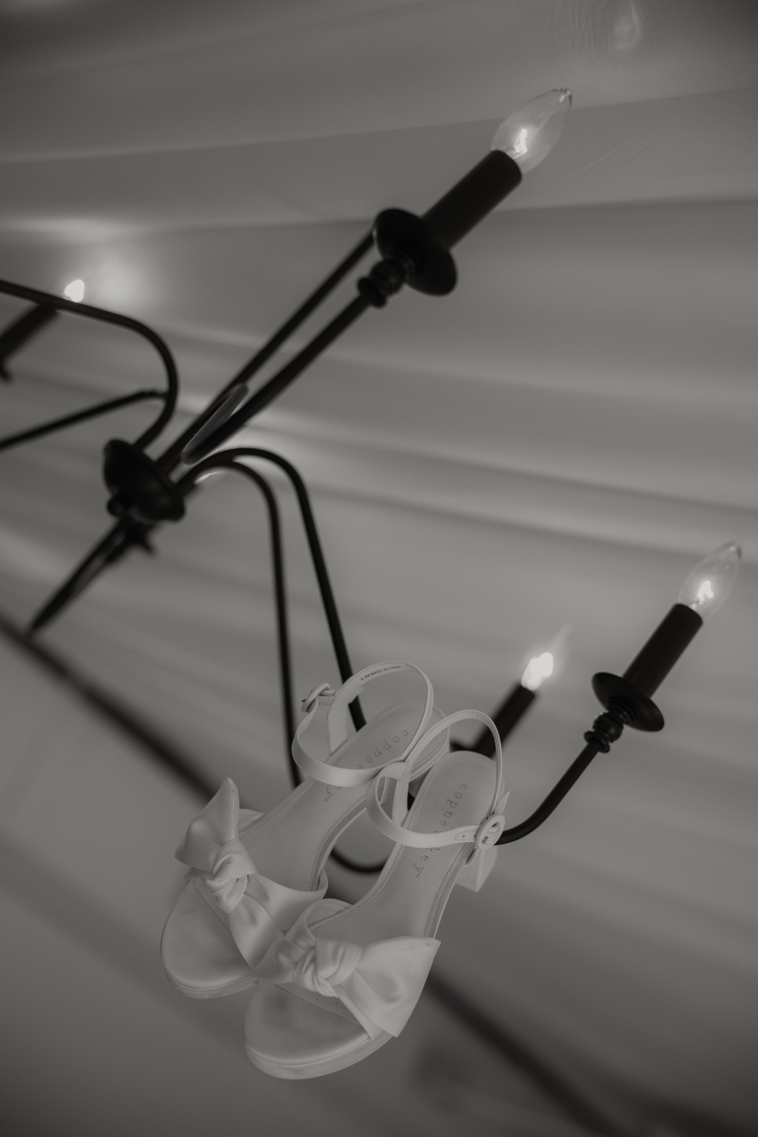White high-heeled shoes with bows hanging from a black chandelier with candle-shaped light bulbs.