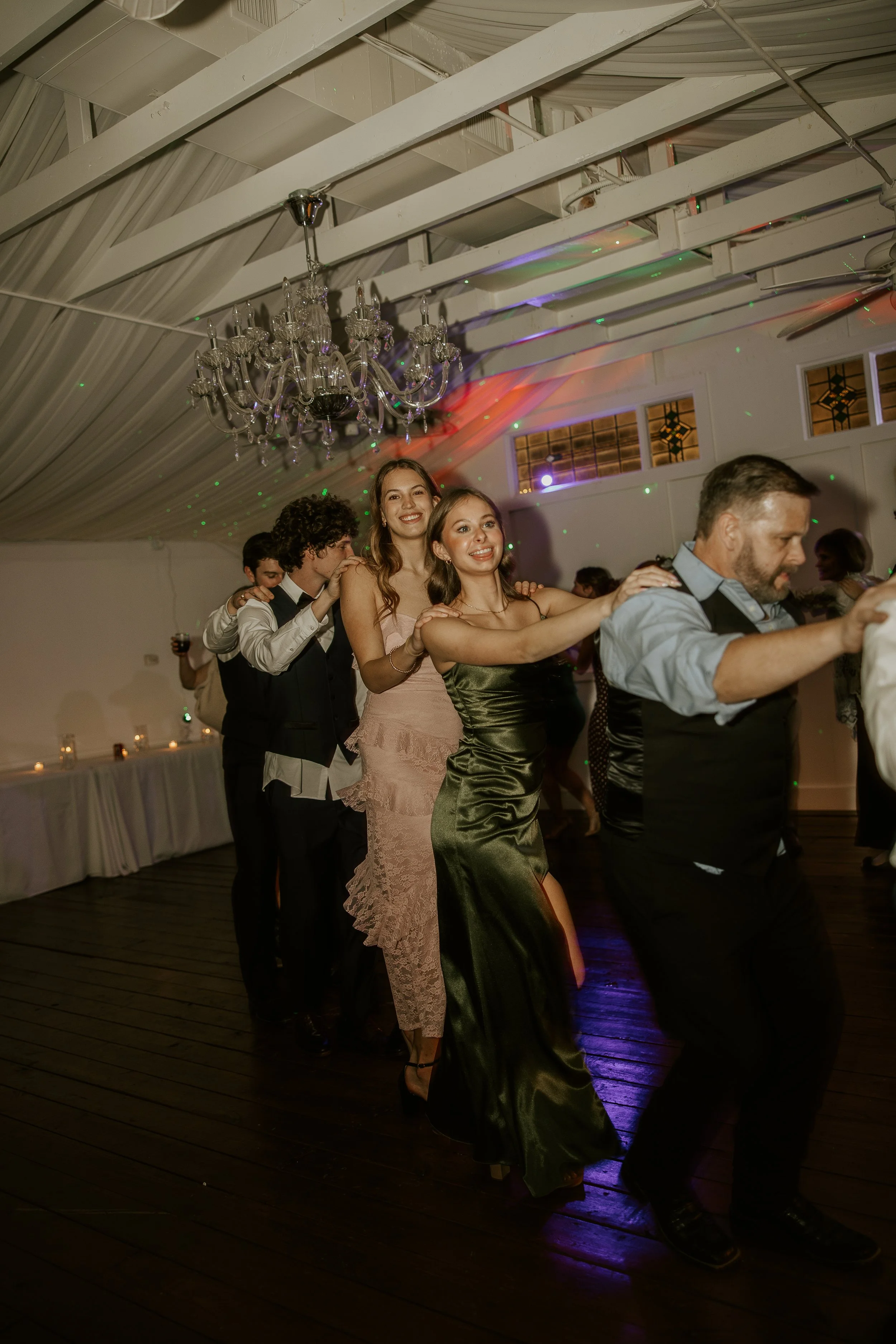People dancing in a line at a celebration, with a chandelier hanging from the ceiling and colorful lights reflecting on the walls.