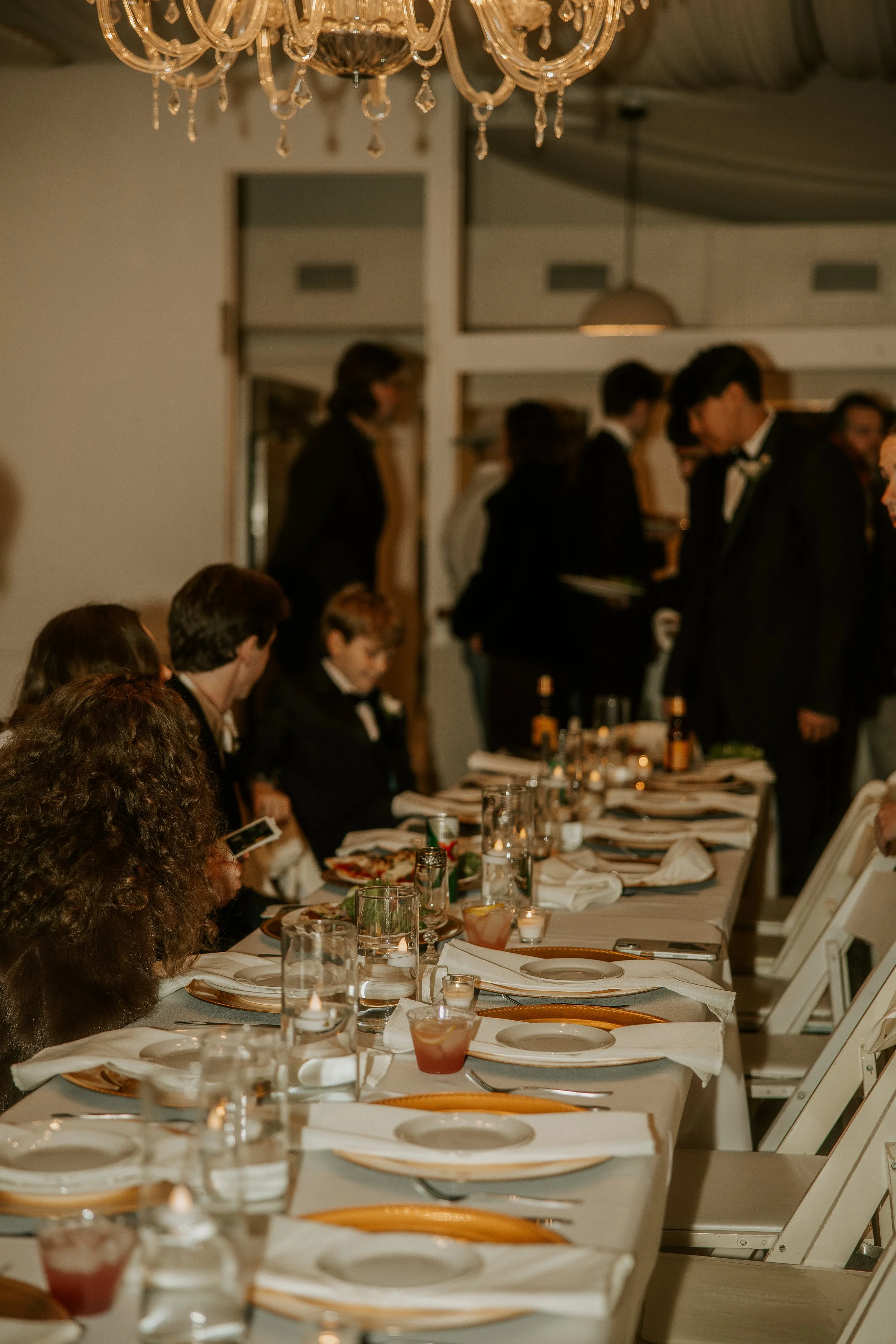 A formal dining setup with a long table, white tablecloth, gold-rimmed plates, glasses, and folded napkins. Several people in tuxedos and formal dresses are seated and standing around the table, with some engaged in conversation. A chandelier hangs a