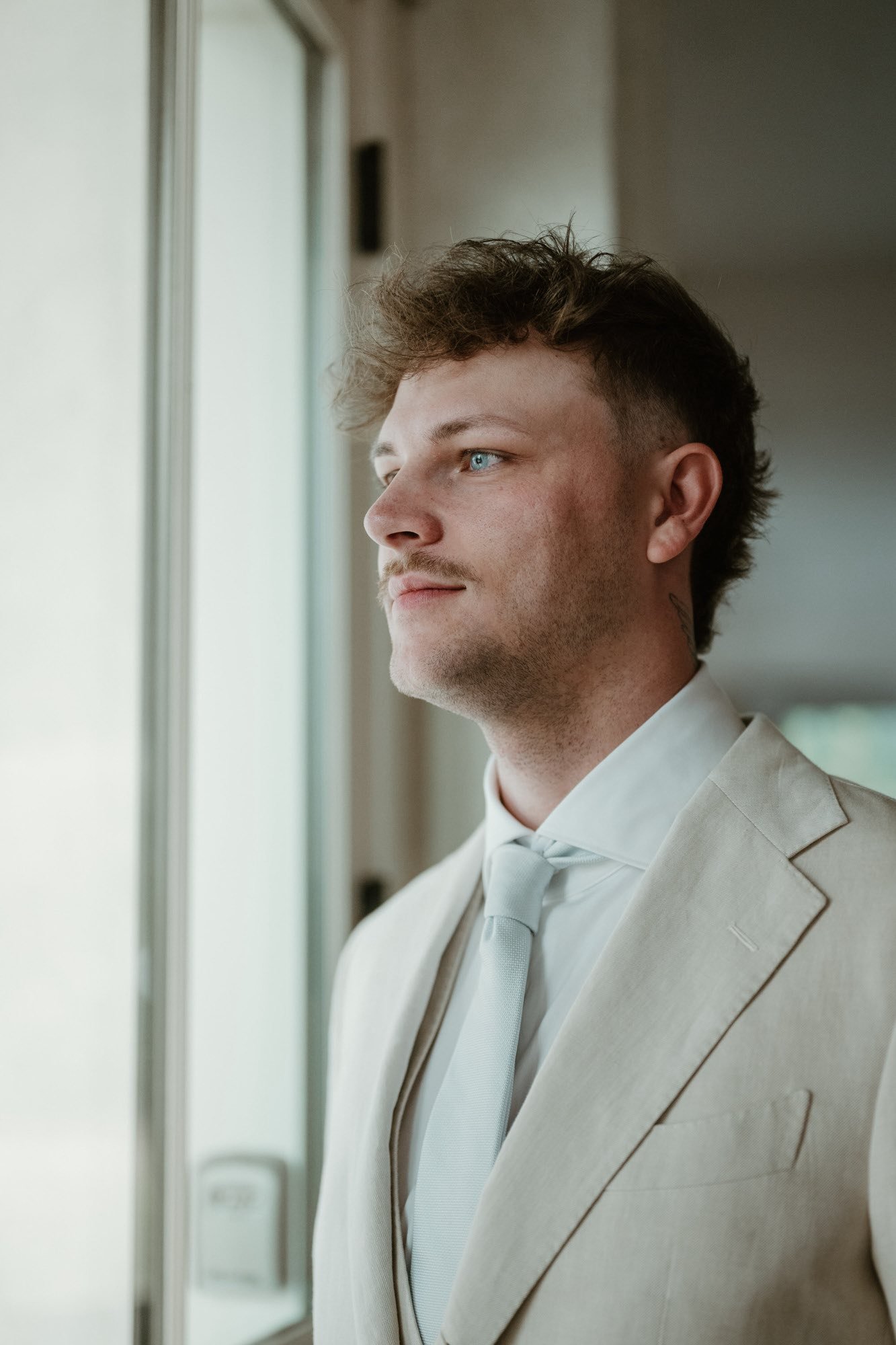 A man dressed in a light-colored suit and tie looking out a window.