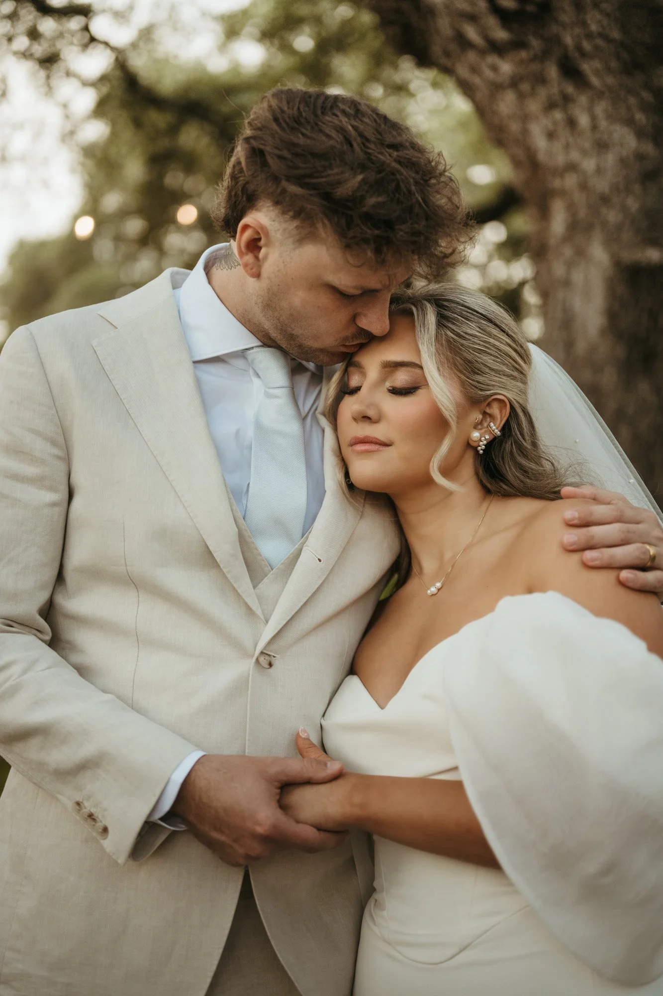 A couple in wedding attire sharing an intimate moment outdoors, with the man gently kissing the woman's forehead and holding her hand.