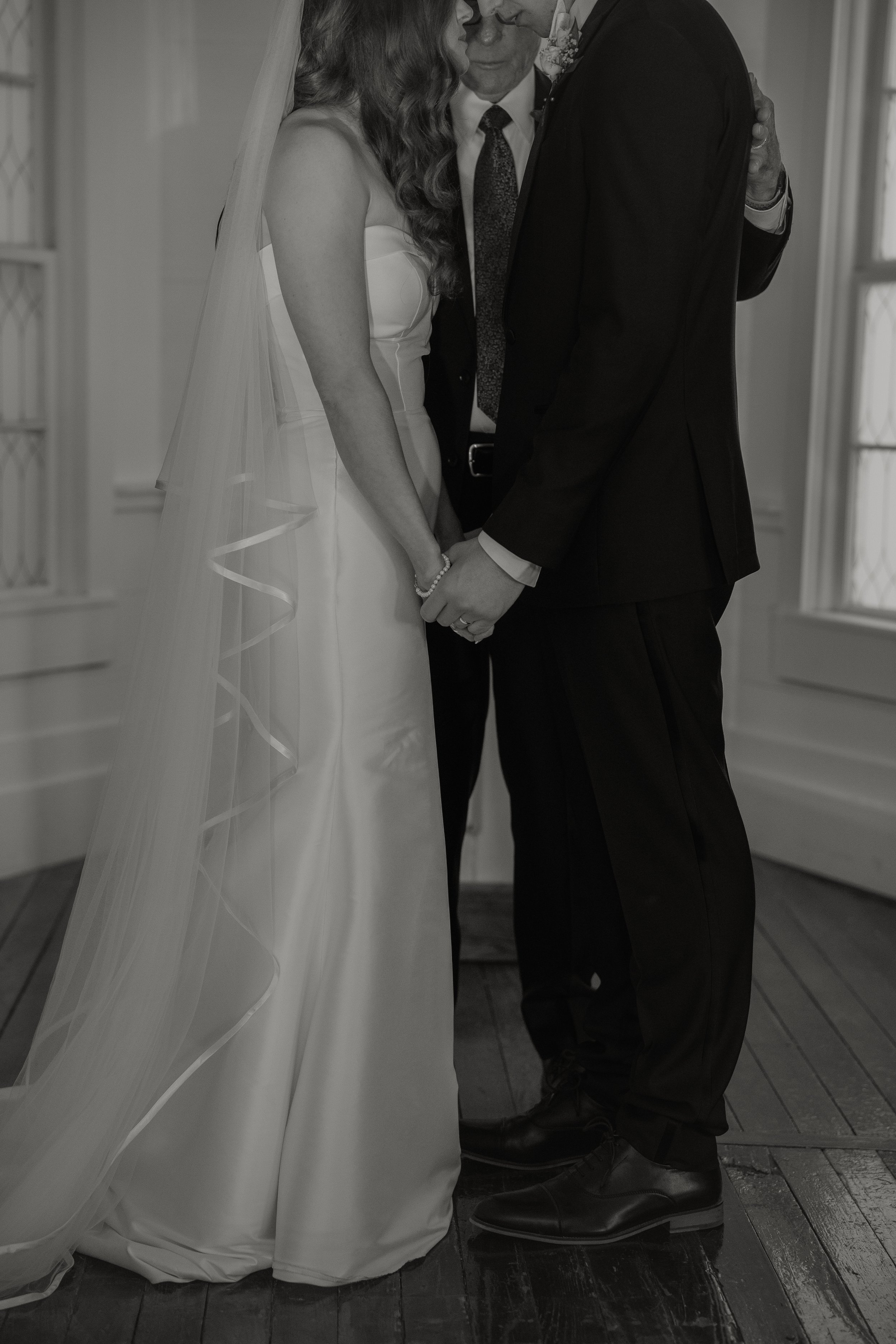 Bride and groom holding hands during their wedding ceremony, with an officiant standing behind them, in a room with large windows.
