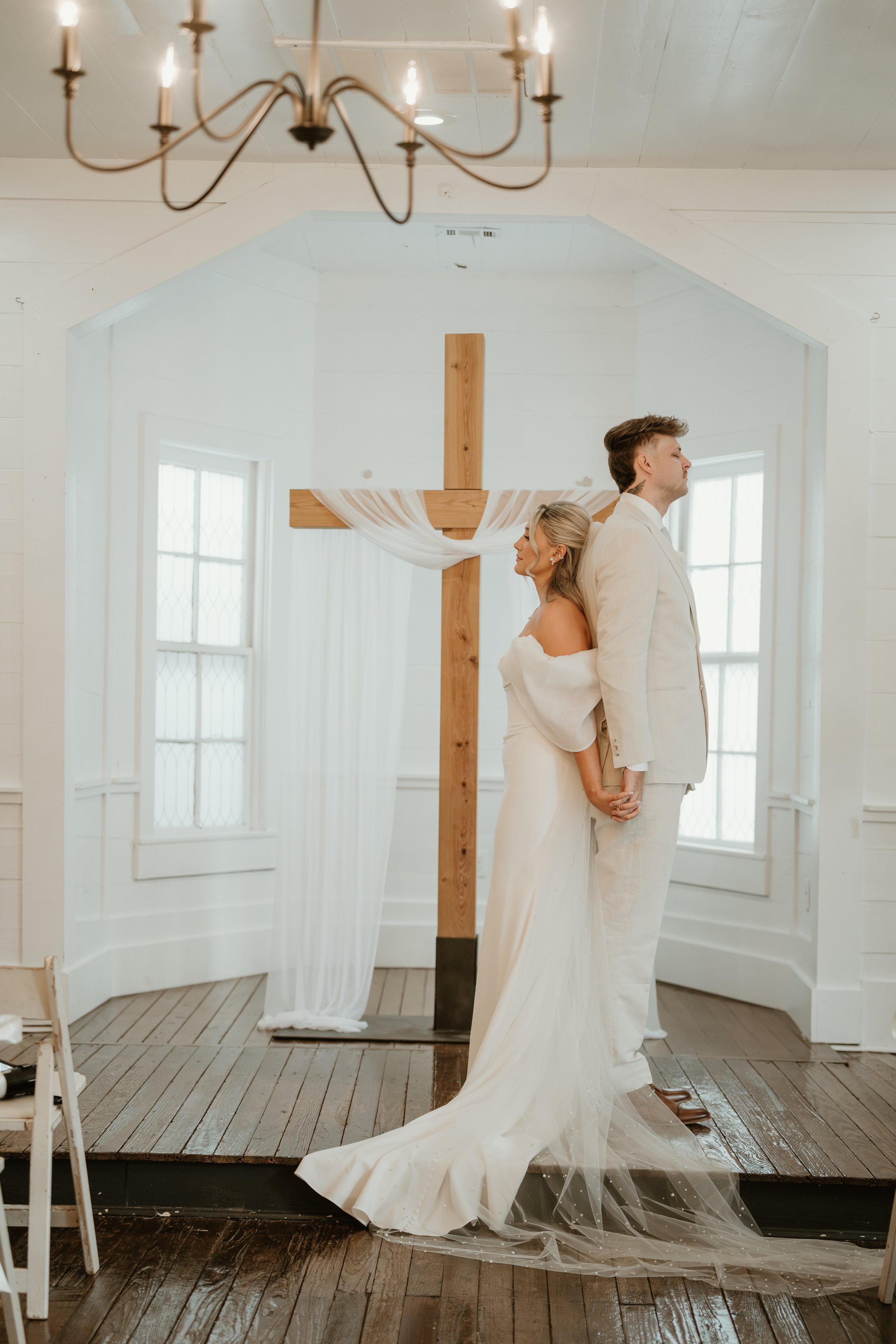 A bride and groom holding hands back-to-back in front of a large wooden cross decorated with white fabric, inside a bright white chapel with wooden floors and windows.