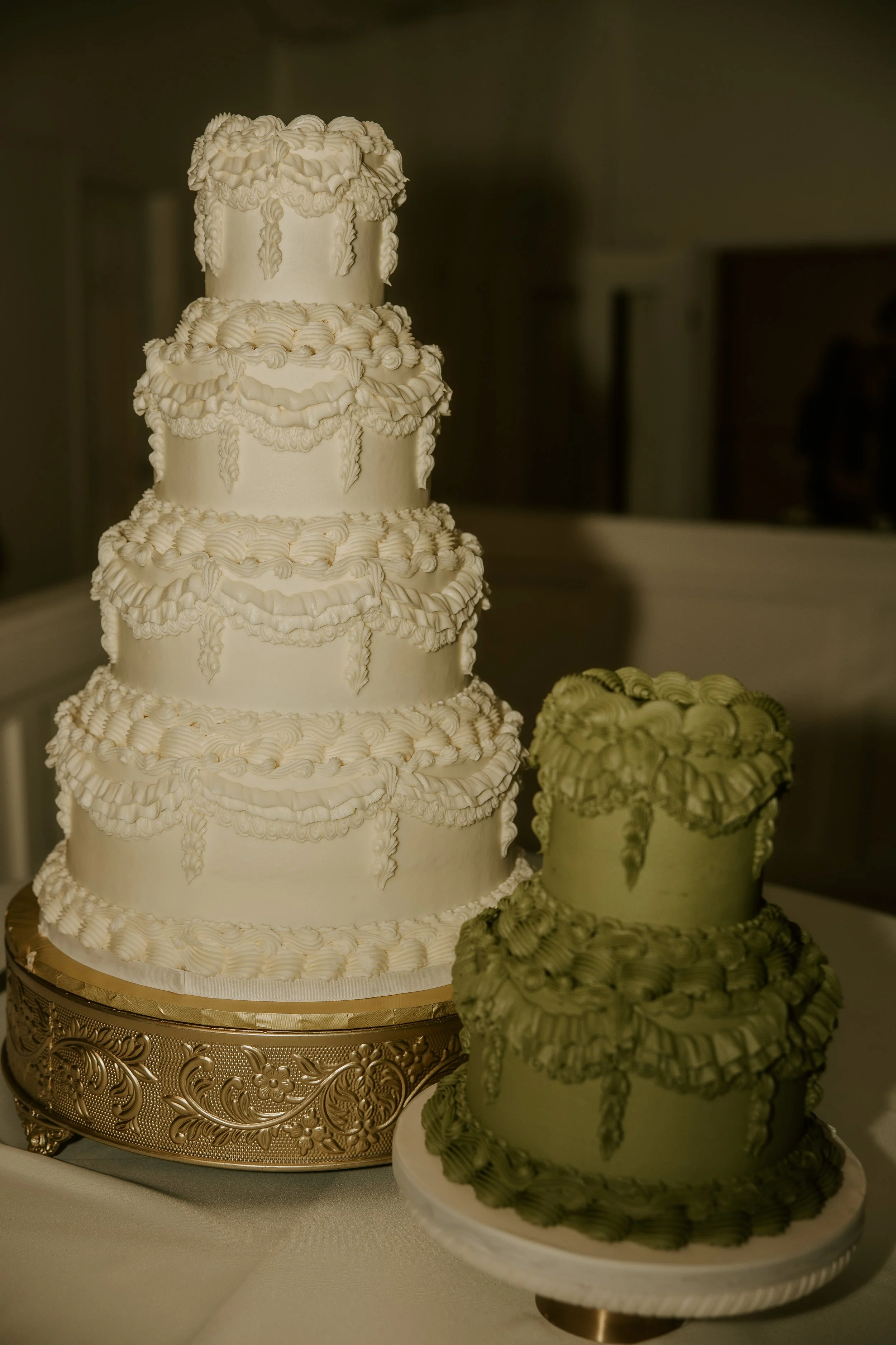 A large, multi-tiered white wedding cake with elaborate piped frosting decorations, placed on a gold decorative cake stand. A smaller, green-toned version of the same cake is next to it.