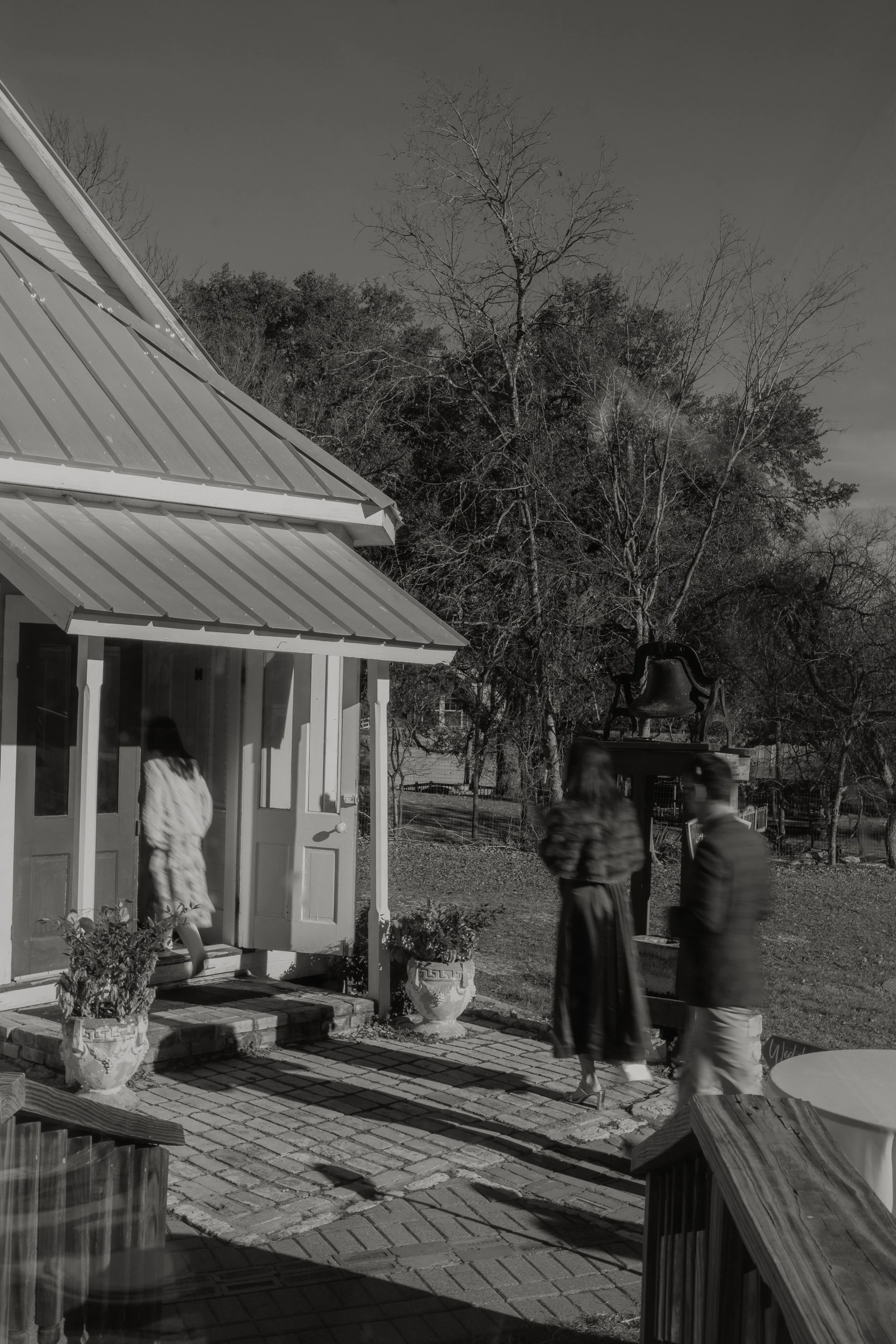 Black and white photo of a house porch with a woman entering, two women and a man walking past, a tree in the background, and a large bell on a stand.