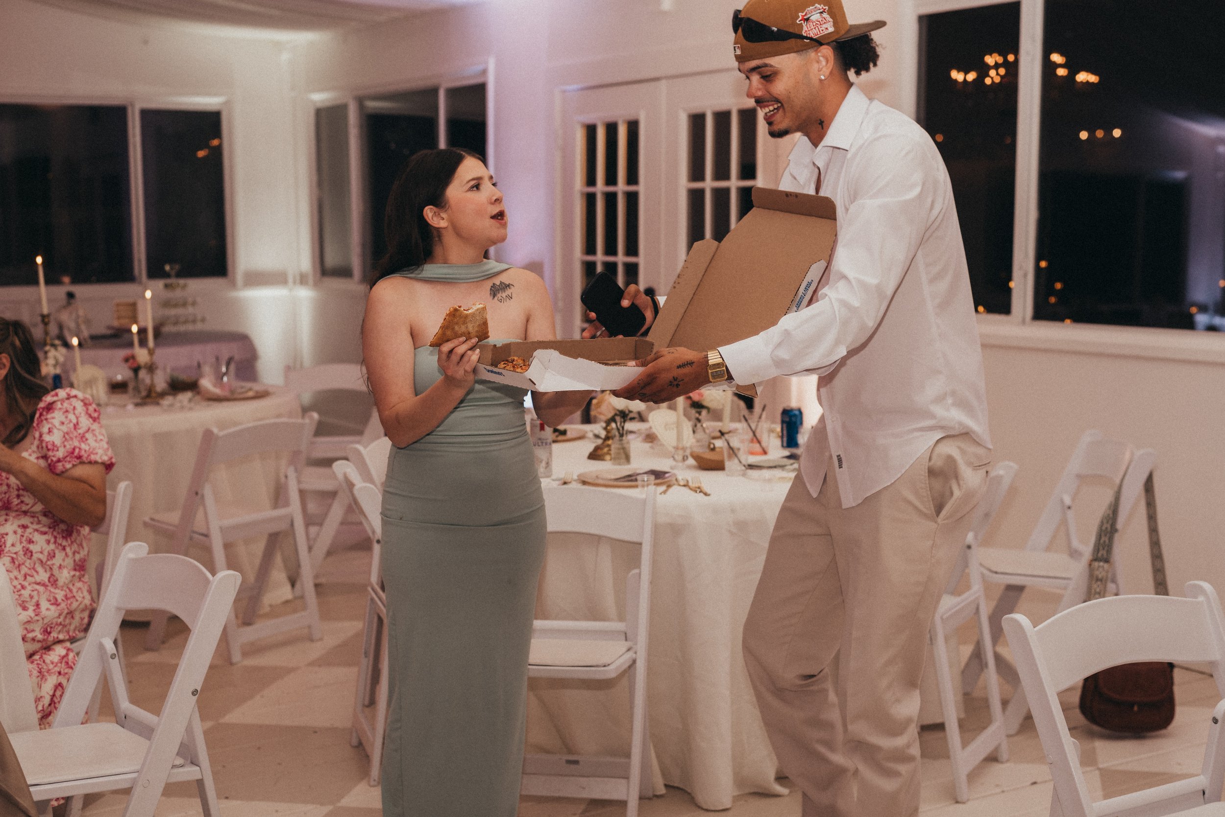 A woman in a gray dress receives a pizza from a man in a white shirt at a social gathering in a decorated indoor venue at night.
