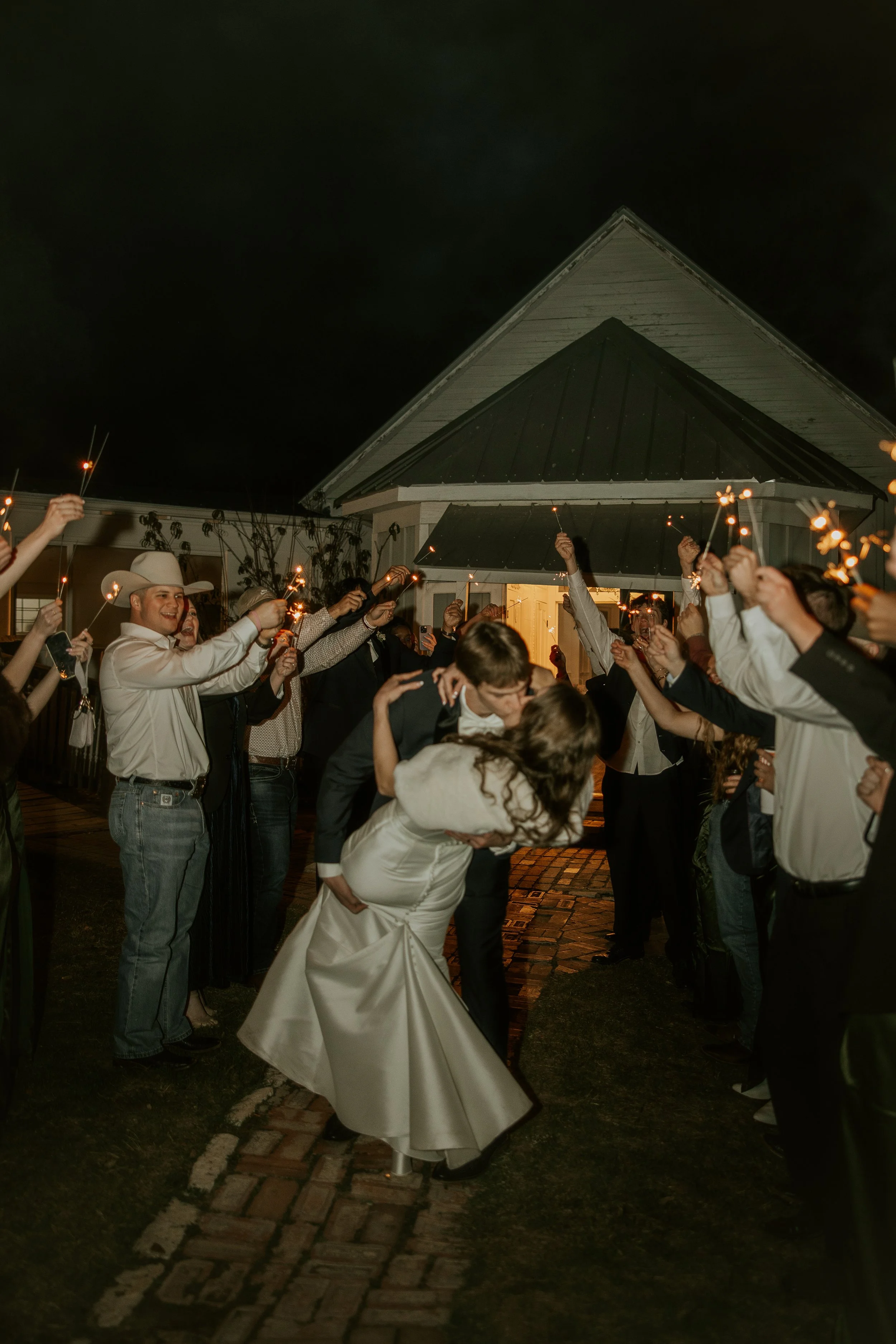 A bride and groom kissing while dancing at their wedding reception outside at night, surrounded by guests holding sparklers.