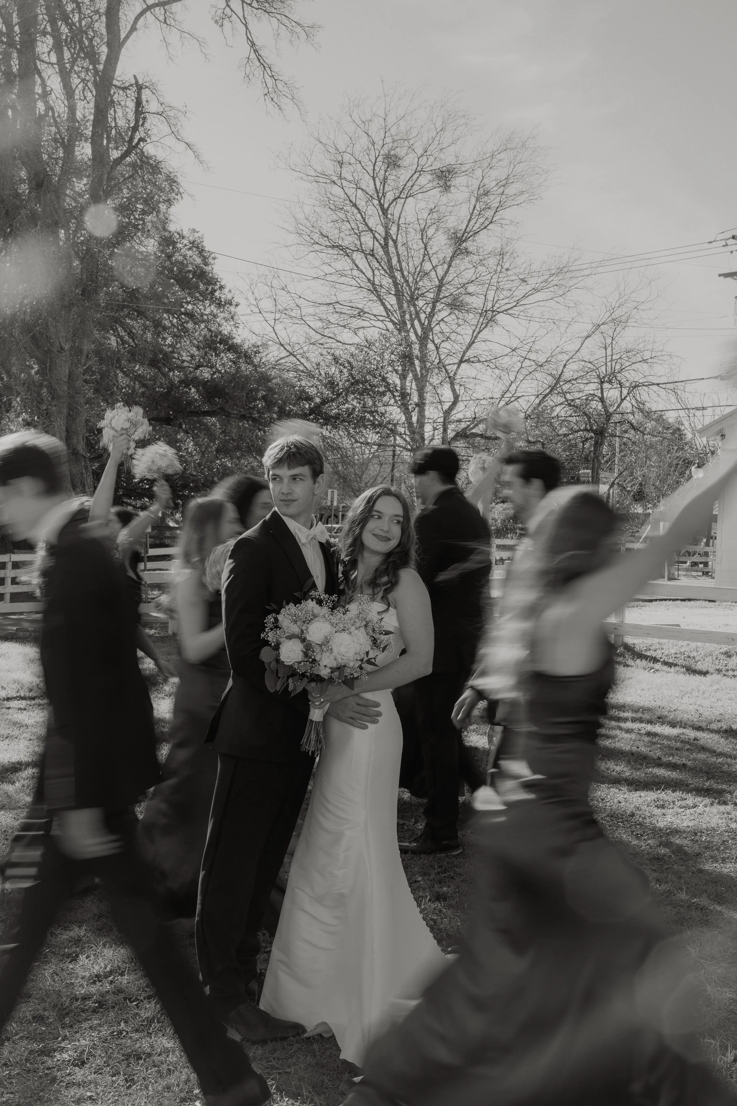 Black and white photo of a bride and groom embracing at a wedding, surrounded by wedding guests walking in a circle outdoors on a sunny day with bare trees in the background.