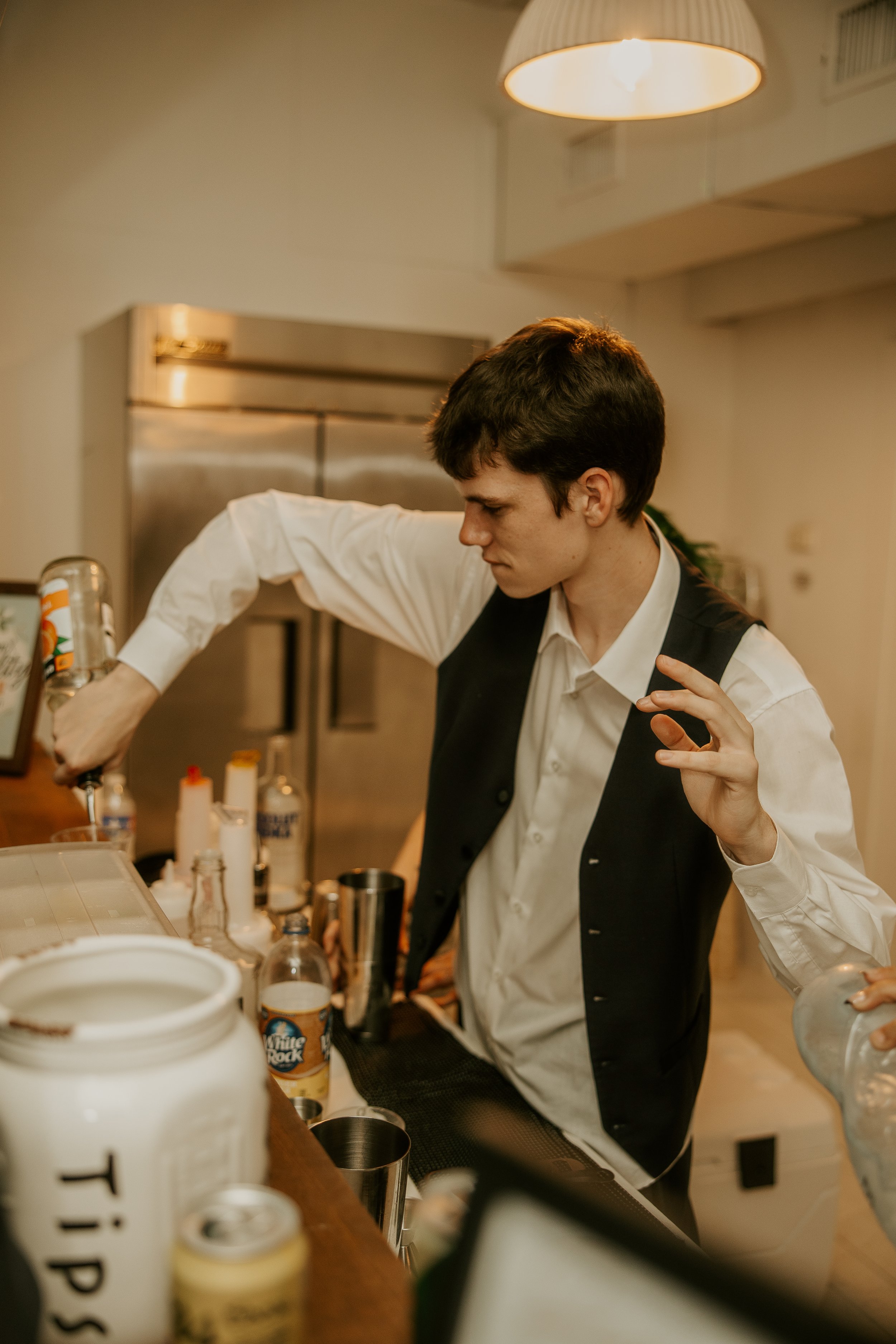 A young man with dark hair dressed in a white shirt and black vest making a cocktail behind a bar, with various bottles and bar tools on the counter.
