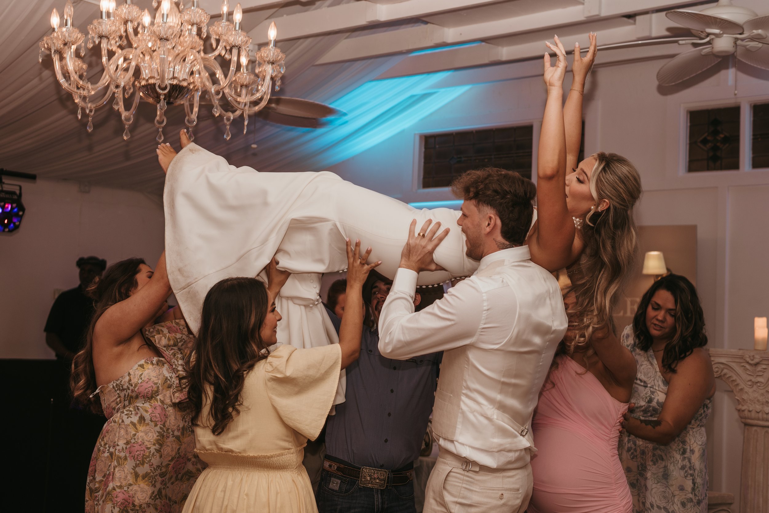 People at a wedding celebration lifting the bride in the air indoors with chandeliers and ceiling fans.