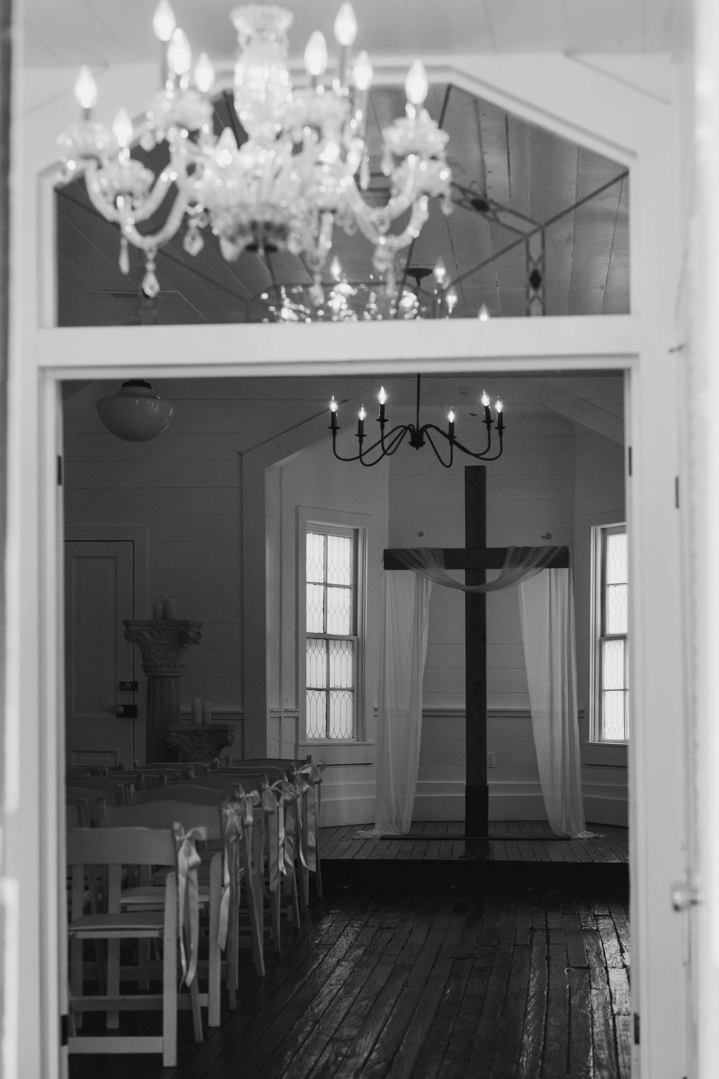 A black and white photo of an interior room with a ceiling chandelier, a wall-mounted chandelier, and a wooden cross with curtains on a small stage, viewed through a window.