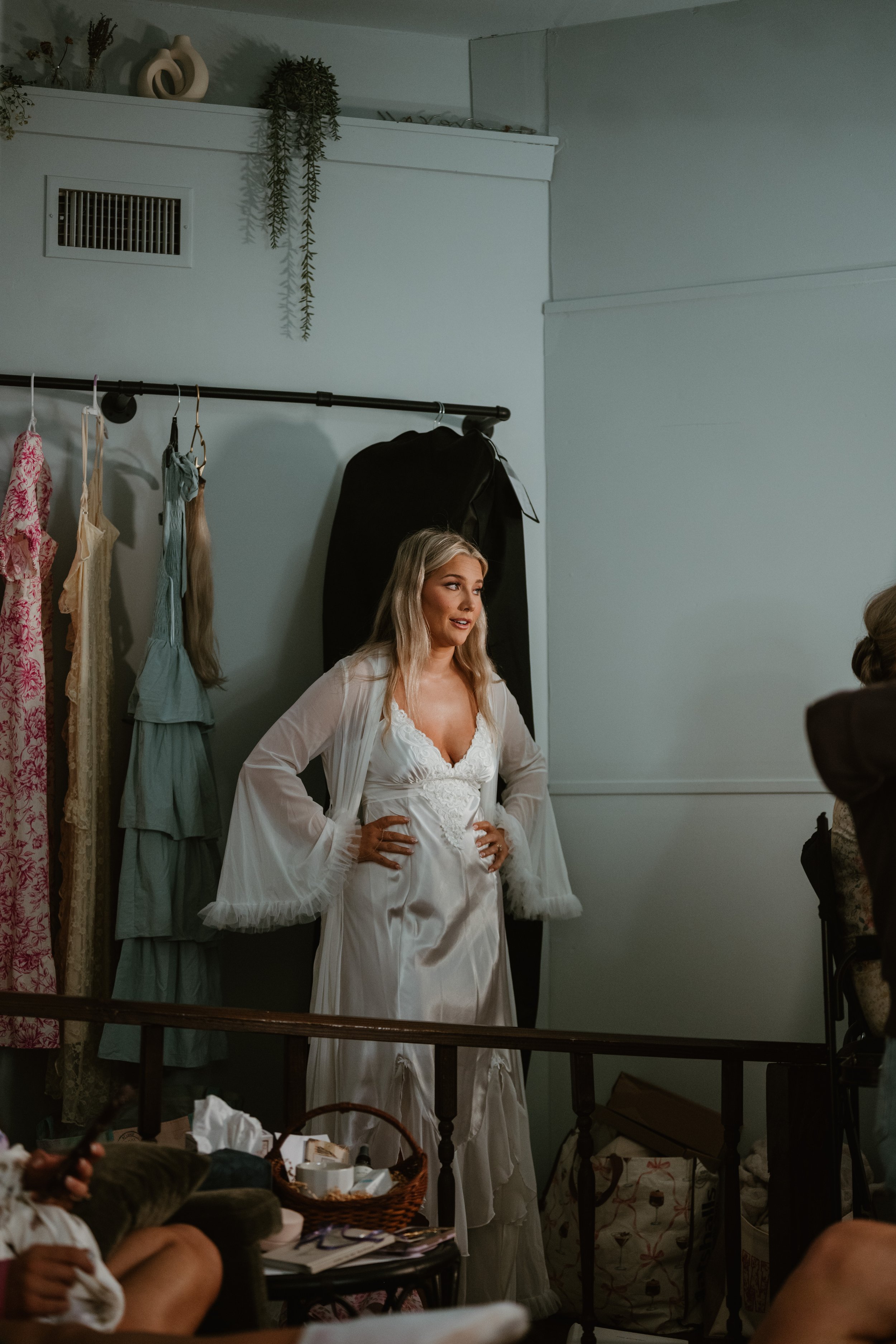 A woman in a white satin or silk lacy slip or robe, which resembles a wedding dress, stands in a room surrounded by dresses and clothing while talking. She has long blonde hair.