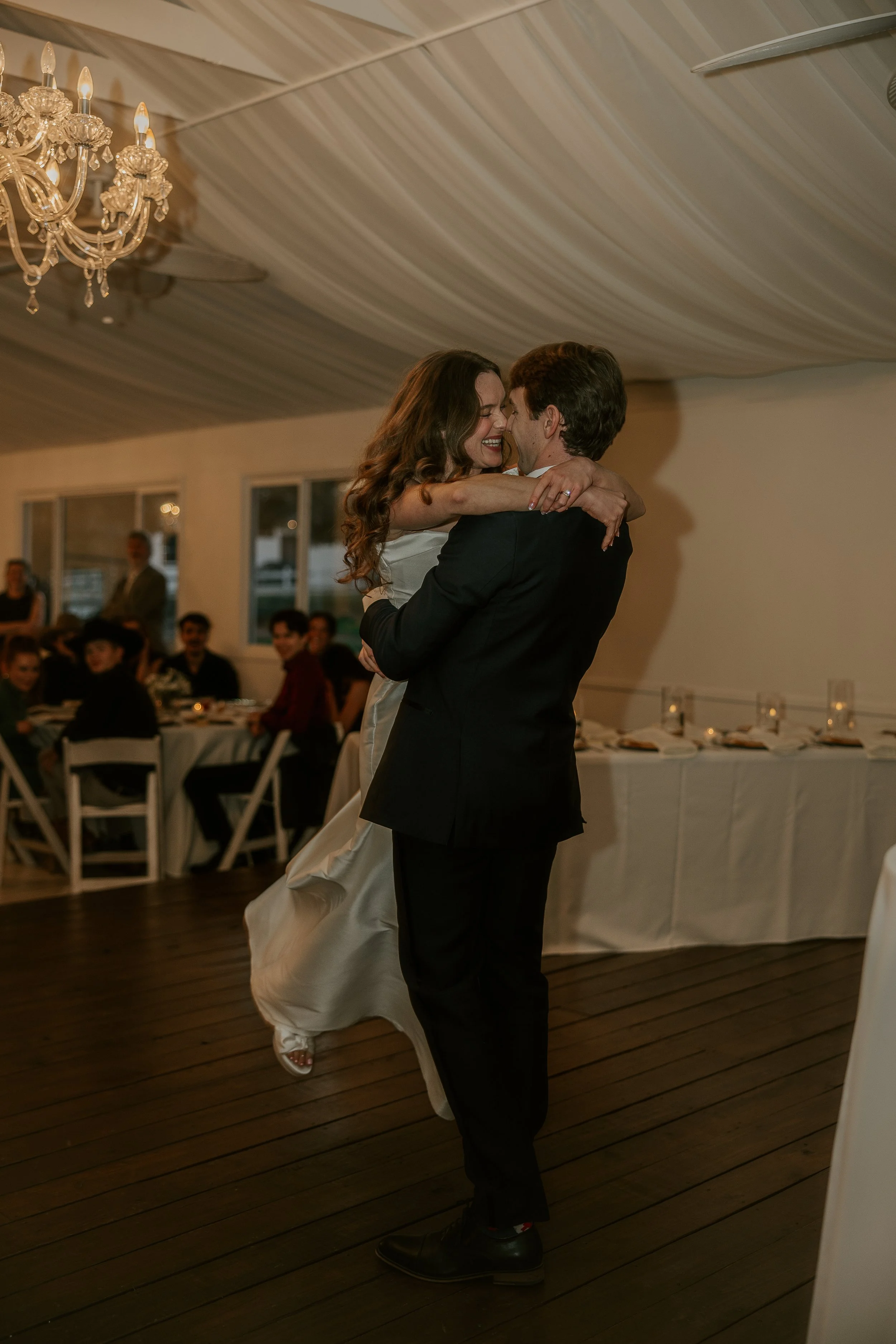A couple dancing and kissing at a wedding reception, with guests seated at tables in the background.