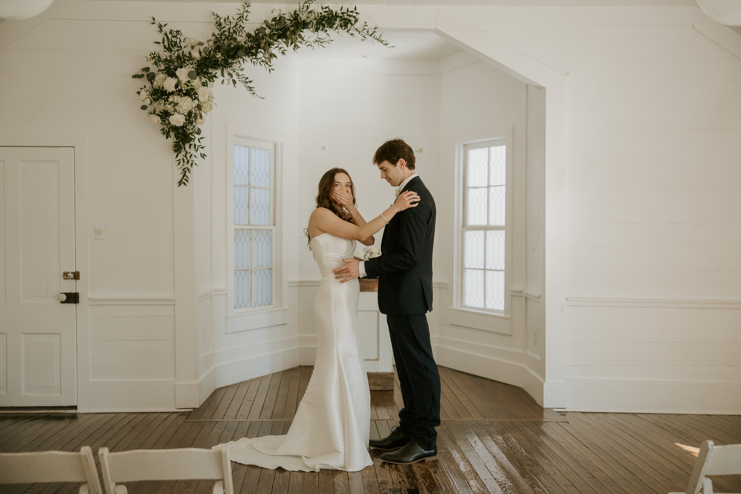 A bride in a white wedding dress and a groom in a black suit share an emotional moment during their wedding ceremony inside a bright, white room with large windows and wooden floors.
