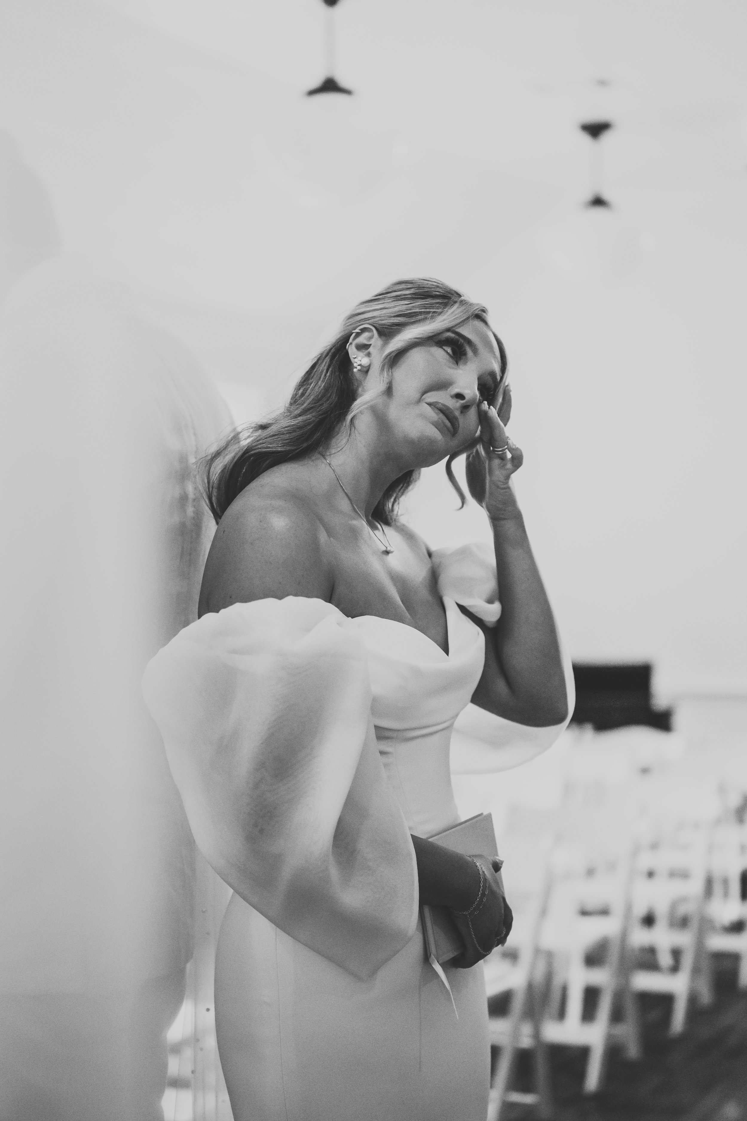A woman in an elegant off-the-shoulder gown standing in a softly lit room, touching her face and appearing emotional.