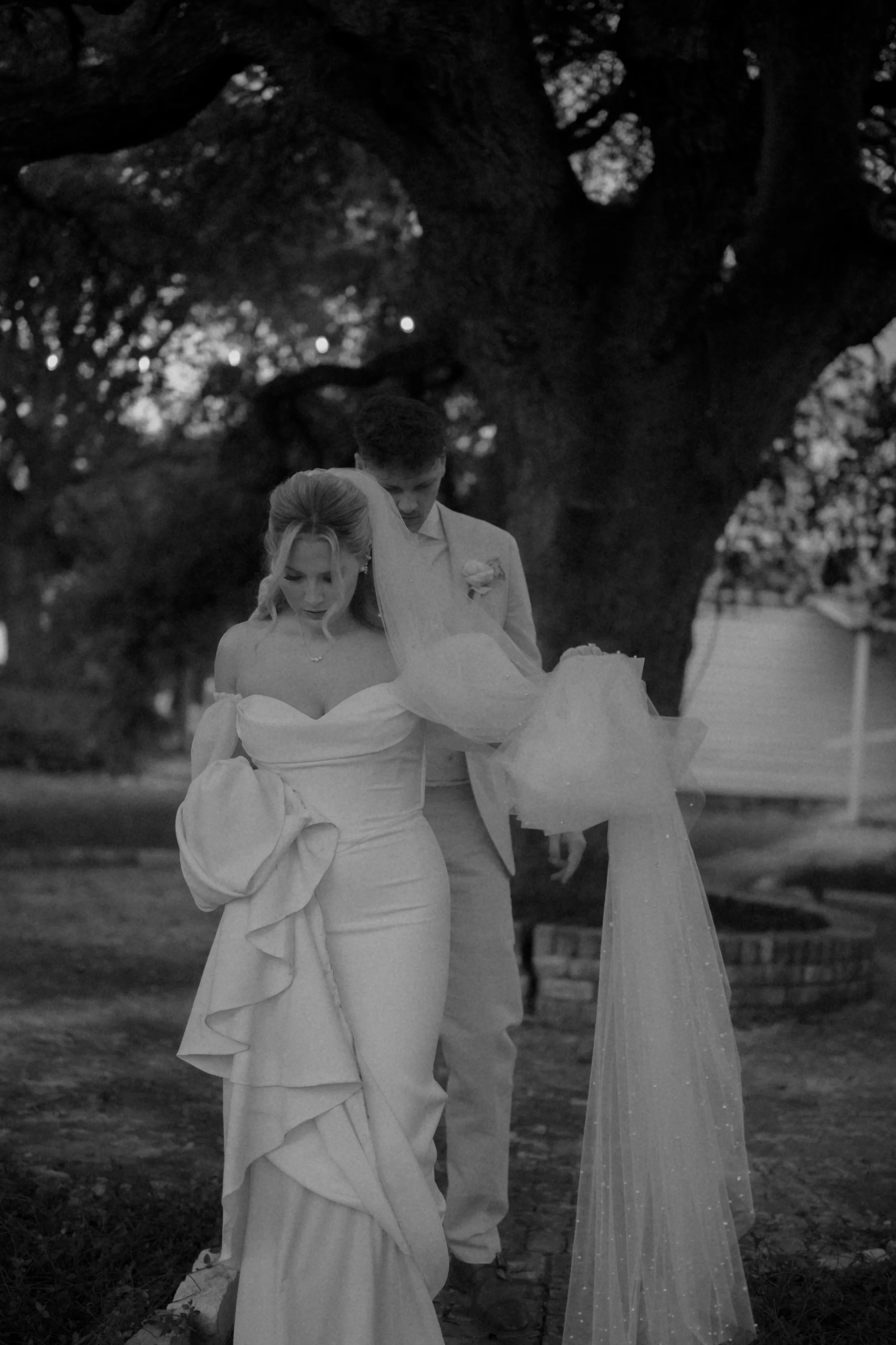 A black and white photo of a bride and groom walking under a large tree during their wedding, with the bride wearing an off-the-shoulder gown and veil, and the groom in a suit, close together.