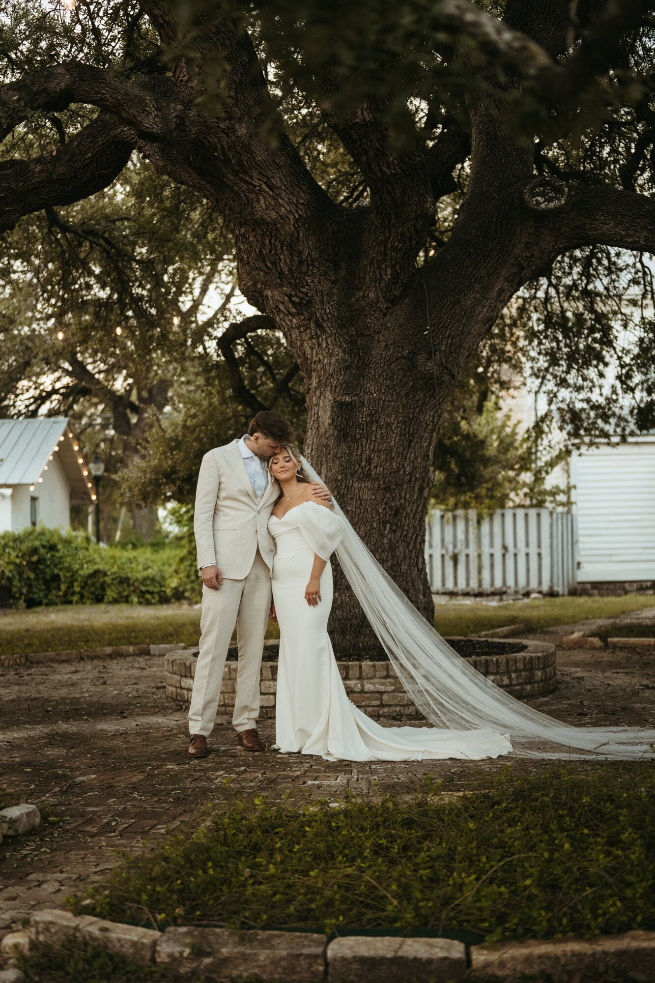 A bride and groom standing close together under a large tree, with the groom embracing the bride and the bride leaning into him, in an outdoor setting during sunset.