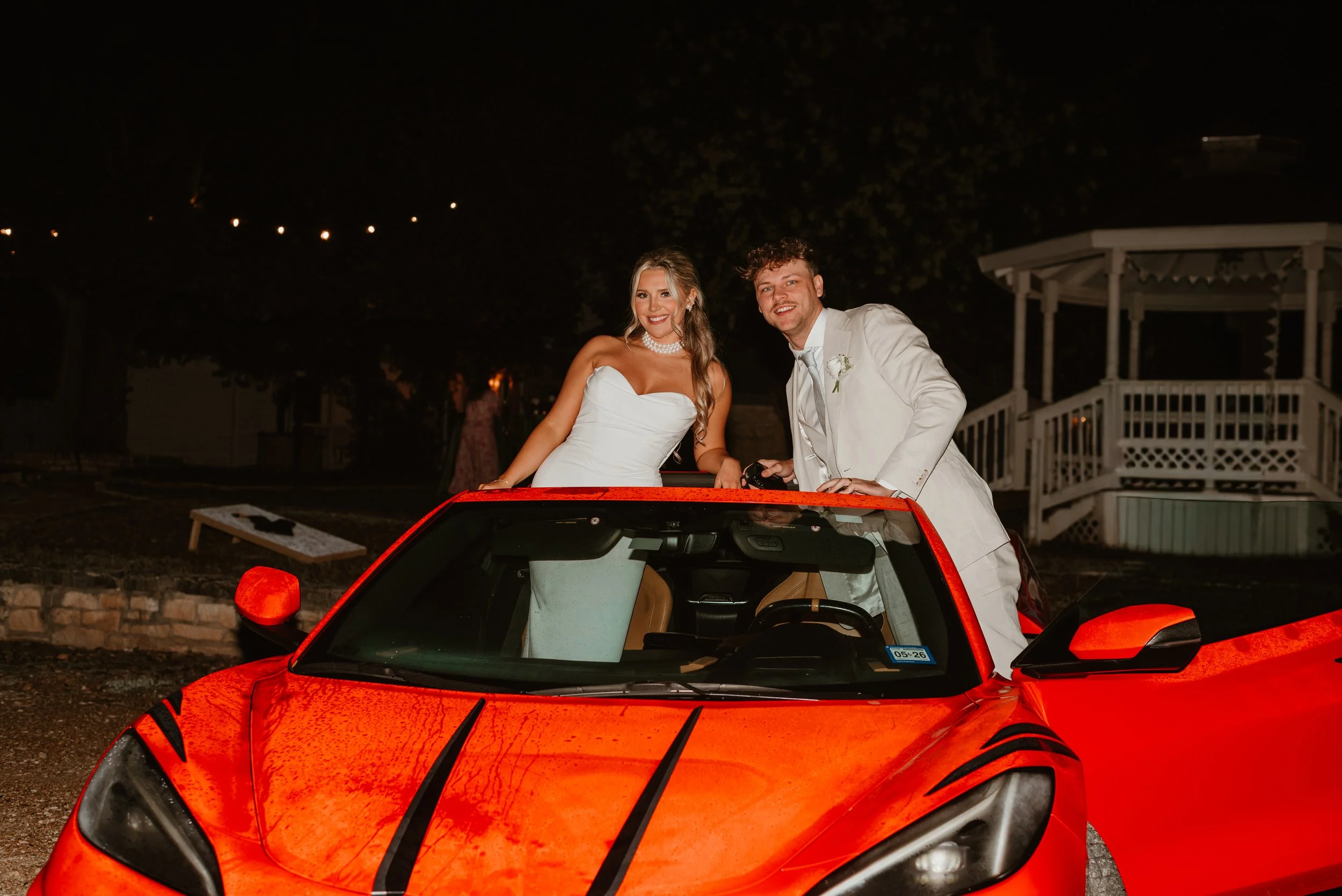 A newlywed couple dressed in wedding attire standing in a red sports car at night, smiling for the camera. The bride is wearing a white strapless wedding gown, and the groom is in a white suit with a boutonniere. The background is dark with some ligh