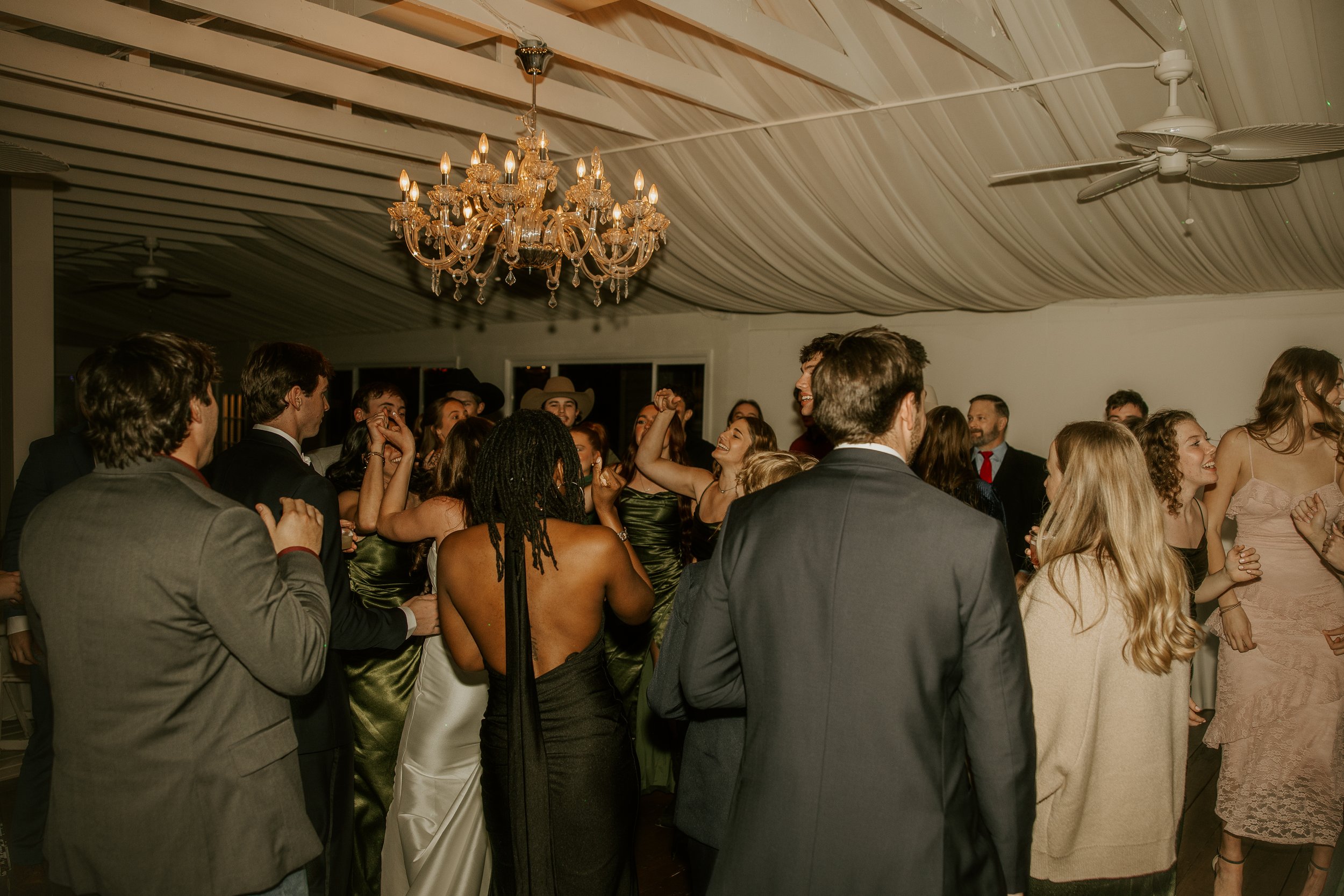People dancing and socializing at an indoor event under a chandelier and ceiling fans.