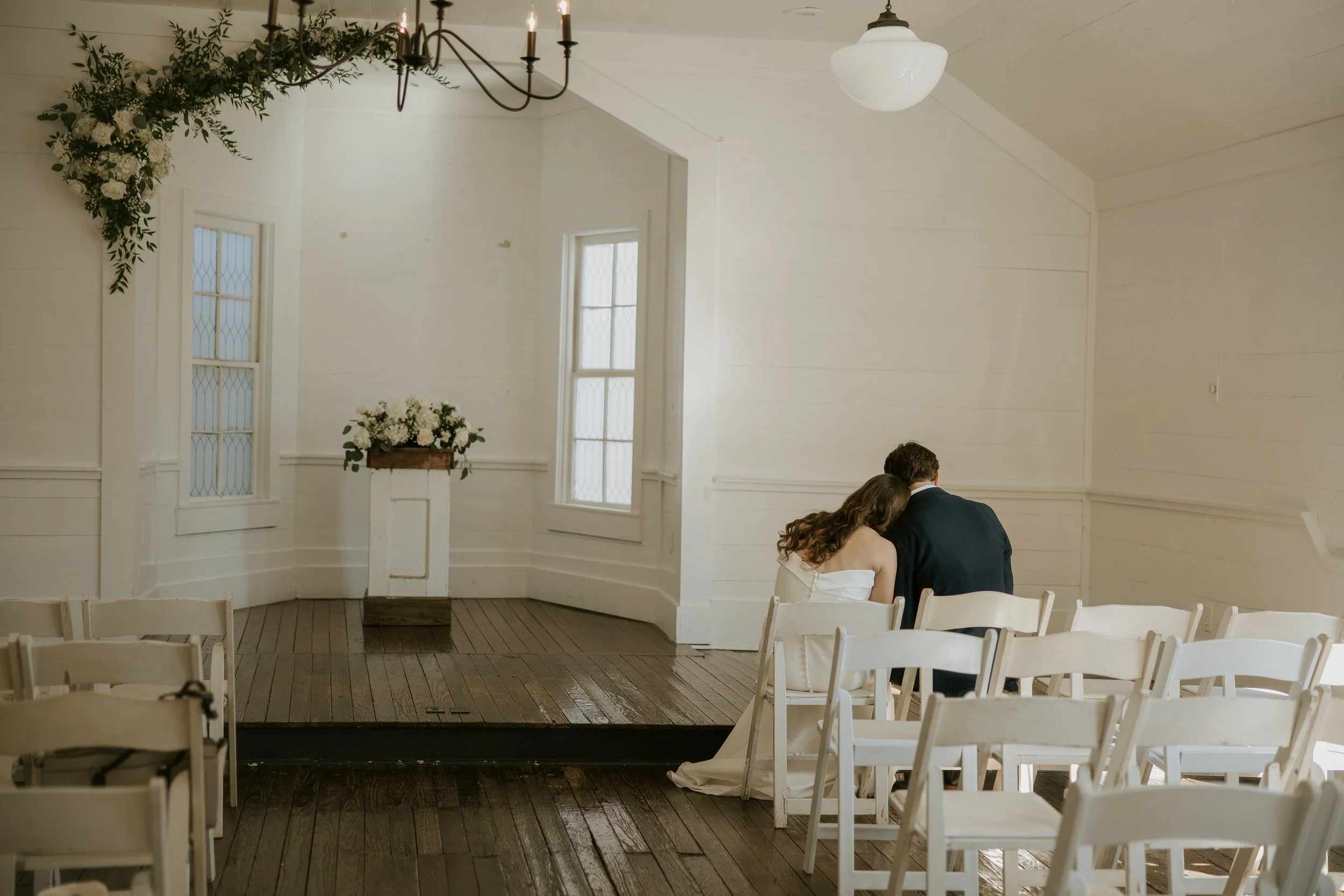 Couple sitting together in a small, bright wedding chapel, with white chairs and floral decorations, likely during a wedding ceremony