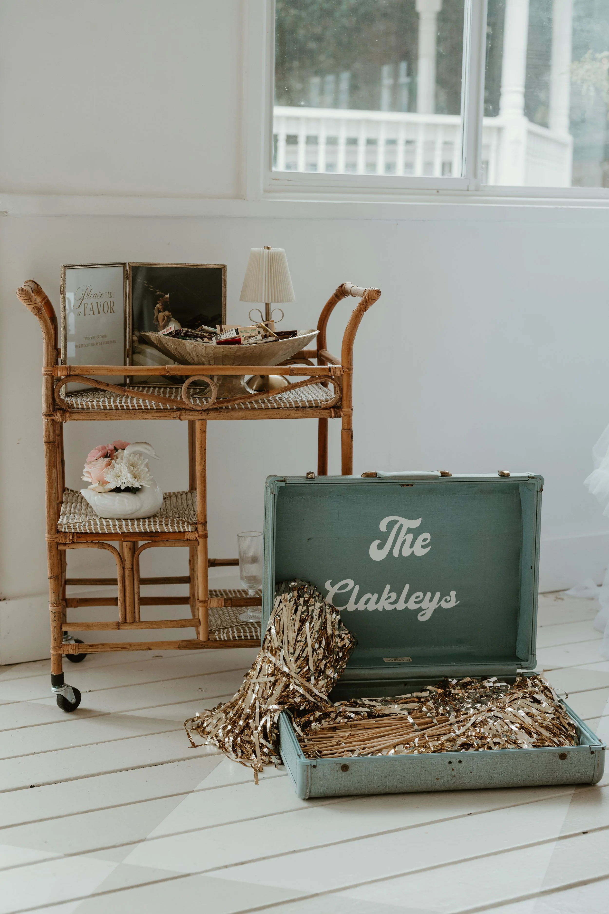 Decorative vintage suitcase with 'The Oakleys' written inside the lid, party decorations, a rattan cart with a lamp, framed photographs, and a small ceramic swan with flowers, set near a window on a white wooden floor.