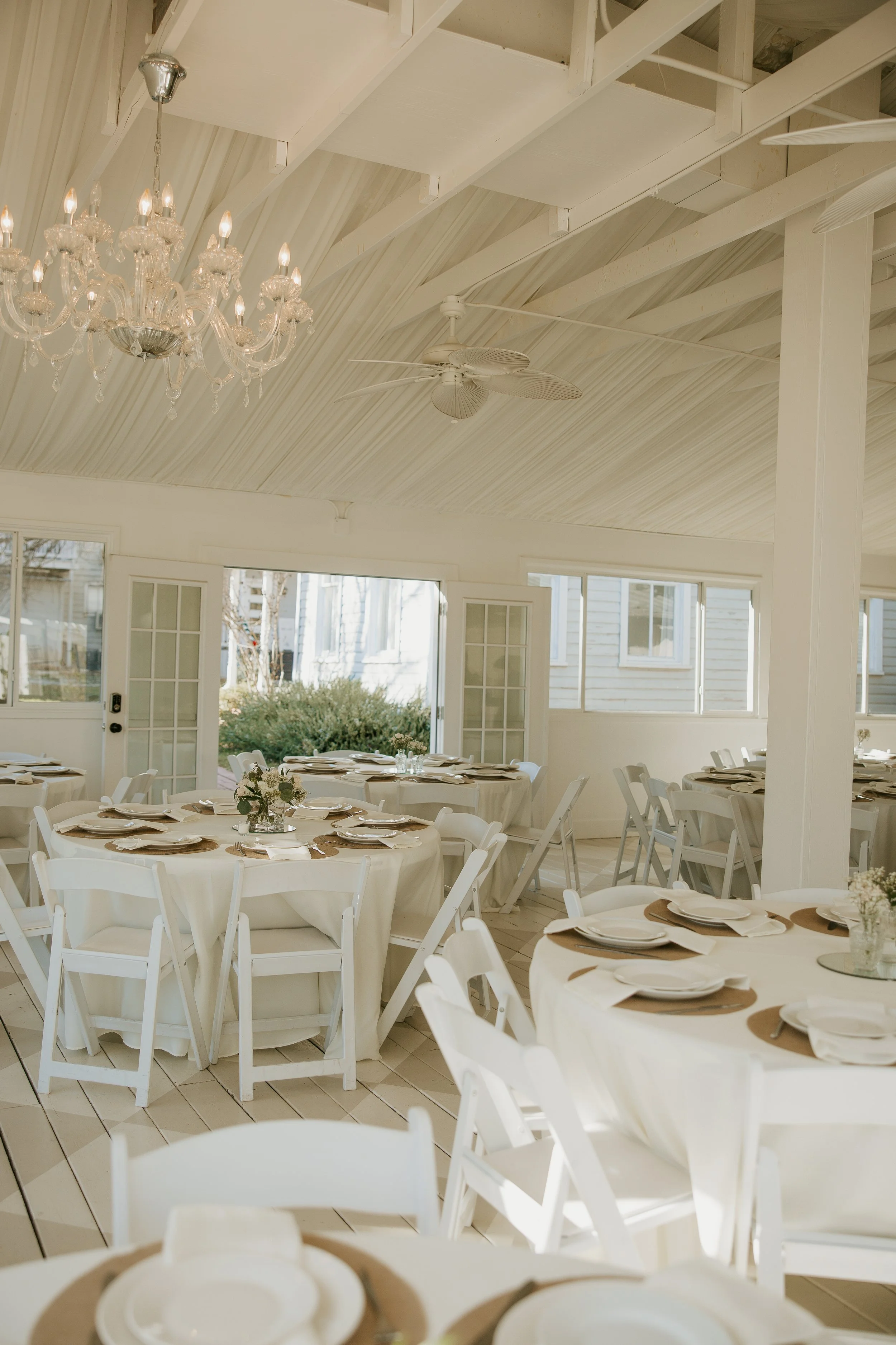 Interior of a bright, elegant dining area with round tables covered in white tablecloths, set with white plates, napkins, and small flower centerpieces. The room features white walls, ceiling beams, a chandelier, a ceiling fan, and open windows letti