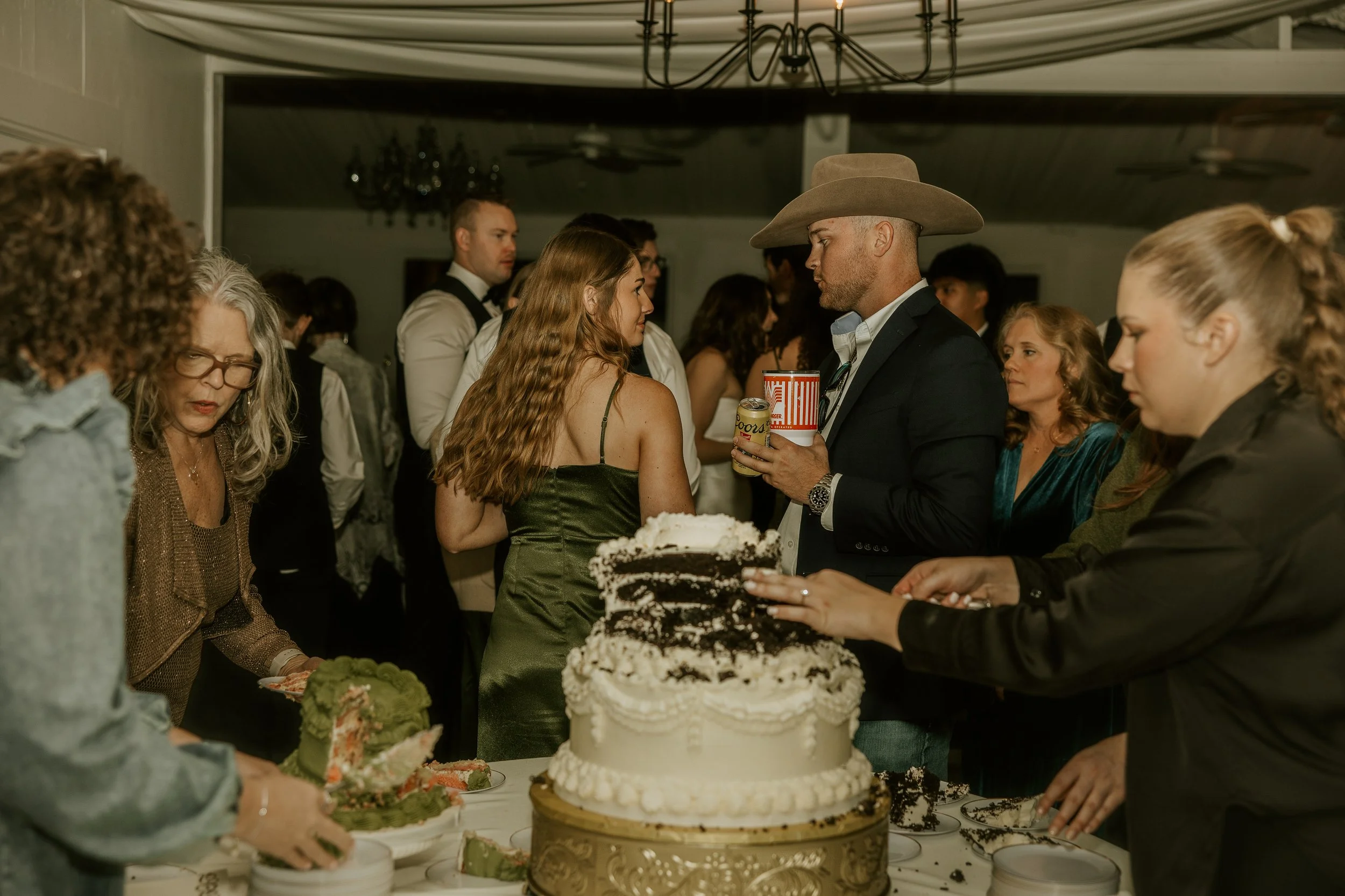 People gathered around a table with a large decorated cake at a celebration party. Some are serving cake, others are conversing, and a man is holding beverages wearing a cowboy hat.