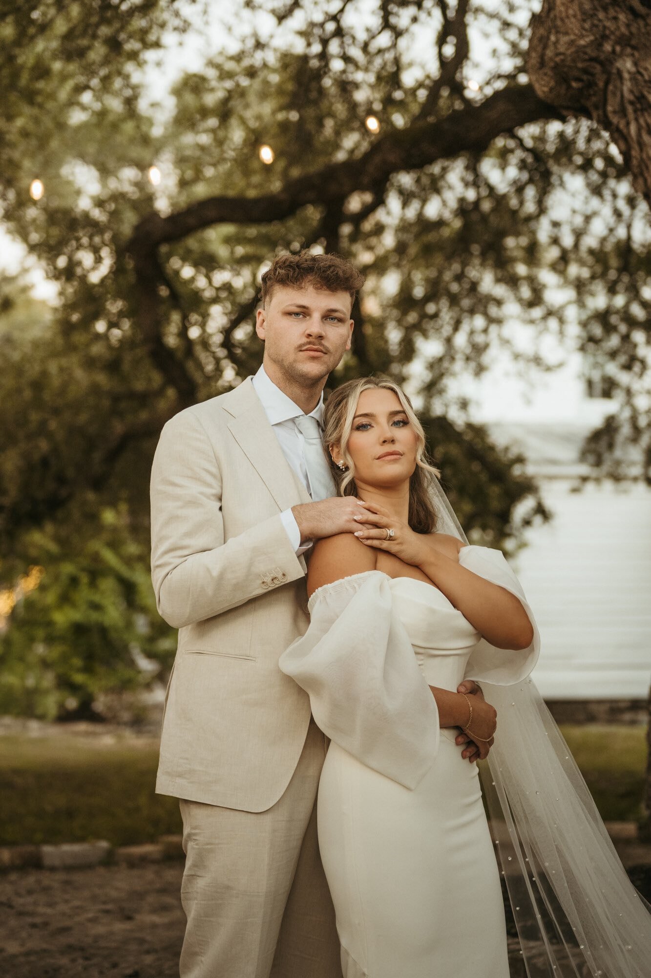Bride and groom posing outdoors, with bride in a white wedding dress and veil, and groom in a beige suit, standing under a large tree with string lights.